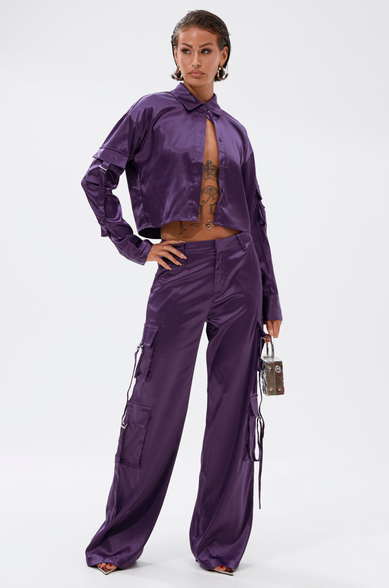 A woman models the AKIRA Label MAYBE ITS YOU SATIN BUTTON DOWN in shiny purple with matching wide-leg cargo pants, accessorized with a metallic handbag and open-toe heels, against a plain white background.