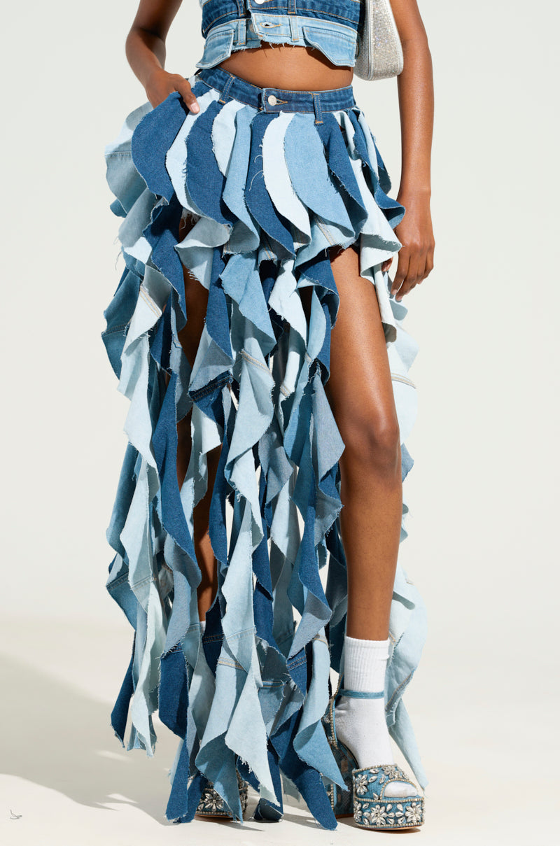 A person wearing the ALESSANDRA DRAMATIC DENIM MAXI SKIRT, designed with a high-rise fit and a button front closure, showcases an outfit of layered, multicolored denim strips. They pair it with white socks and platform sandals against a plain, light-colored background.