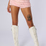 A person standing against a plain background wearing the MADE FOR ME RUFFLE SHORT in pink paired with a matching sheer mesh top, white knee-high lace-up boots, and featuring a noticeable tattoo of a skull on their right thigh.