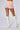 A person wearing the MADE FOR ME RUFFLE SHORT in a high-rise pink design and white high-heeled boots faces away from the camera, showcasing tattoos of crossed arrows on the back of their thighs. The backdrop is a plain white canvas.