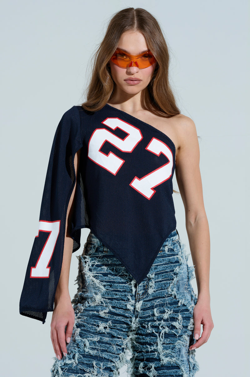 Wearing a sporty-chic ensemble, the person models orange sunglasses and an AKIRA Label black one-shoulder GAME TIME JERSEY with a white and red number 27. The top's sleeve drapes casually alongside distressed blue jeans against a neutral background. 0