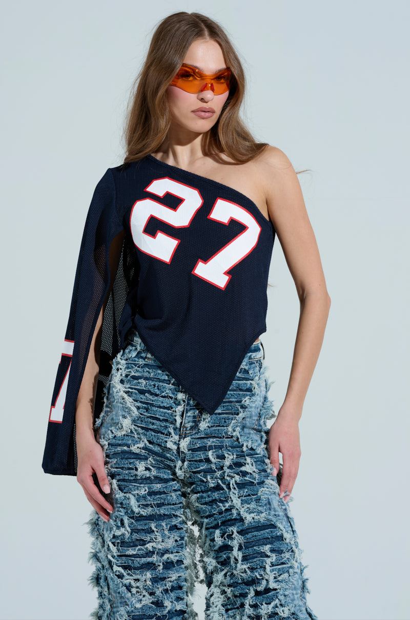 Against a light blue backdrop, a woman showcases sporty-chic style in an AKIRA Label GAME TIME JERSEY with the number 27 and distressed blue jeans. She wears orange sunglasses, and her long brown hair cascades over one shoulder. 1