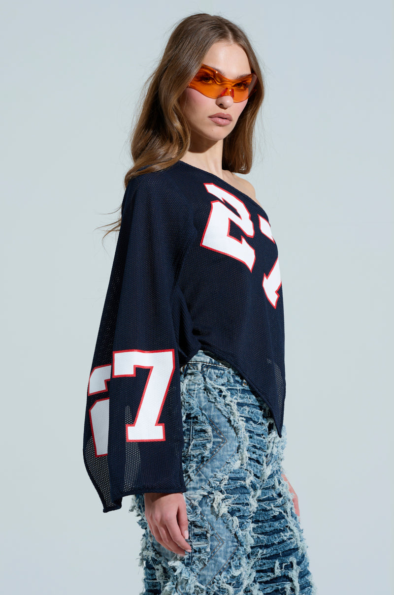 A woman embodies sporty-chic vibes in orange sunglasses and the AKIRA Label GAME TIME JERSEY, showcasing number 27 in white and red. Her blue jeans have a frayed texture on a light gray backdrop, creating a dynamic and stylish look. 2