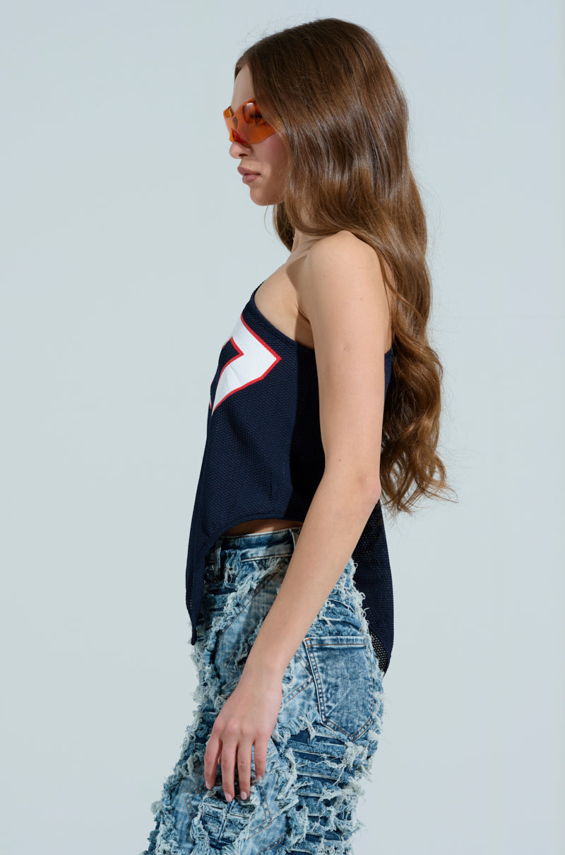 A long-haired woman showcases a sporty-chic look with orange sunglasses and a black sleeveless top akin to the GAME TIME JERSEY, featuring white and red accents. Her distressed blue denim pants add edge against the plain light gray backdrop. 3