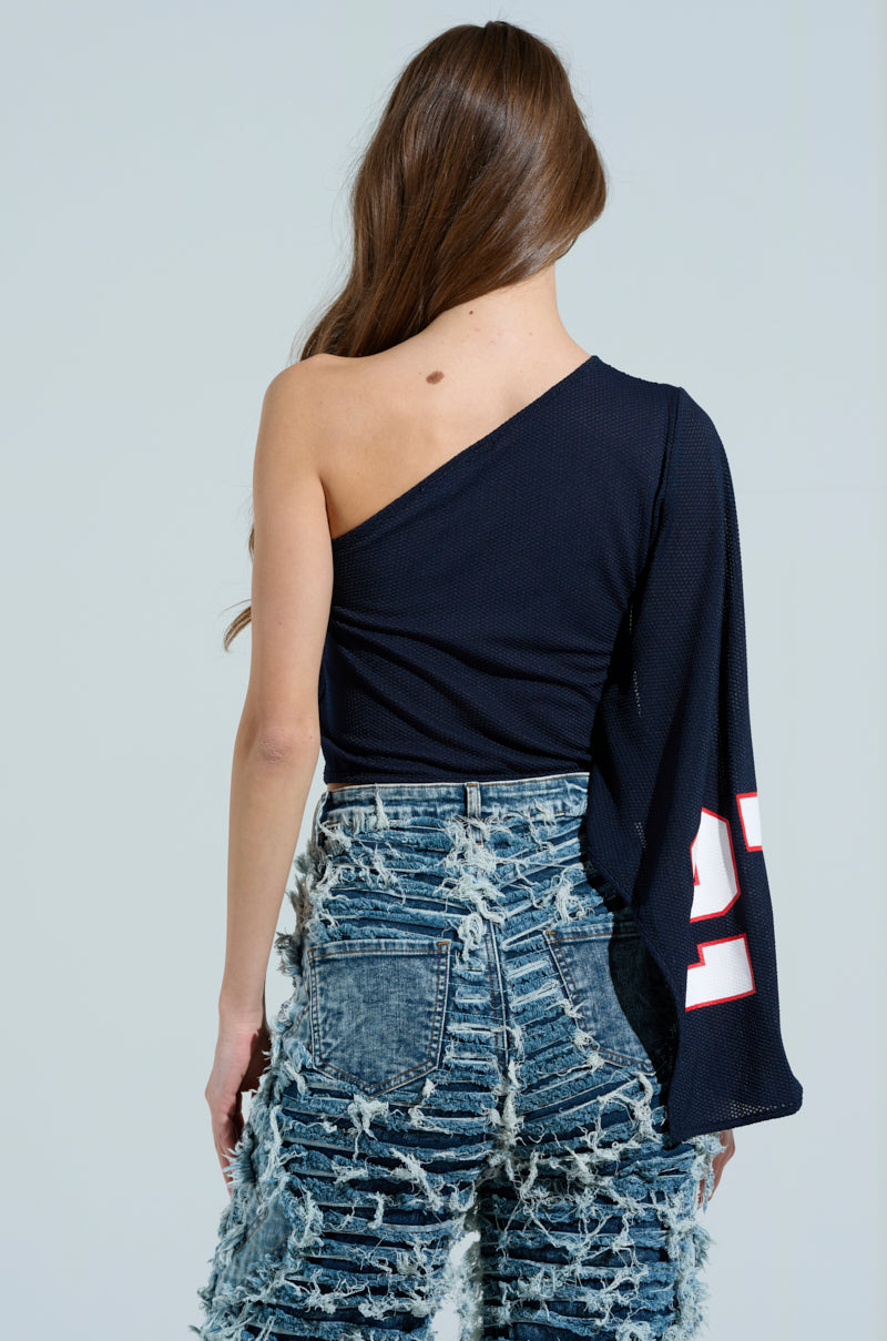 A woman with long brown hair faces away, exuding a sporty-chic vibe. She's wearing the AKIRA Label GAME TIME JERSEY, a dark blue one-shoulder top featuring a bold white number on the sleeve, paired with heavily distressed light blue jeans against a plain gray backdrop. 4