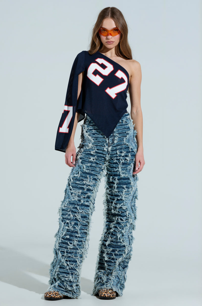 Against a light blue background, a woman poses stylishly in the GAME TIME JERSEY, which is asymmetrical and sports the number 27. Her look is completed with frayed denim bell-bottom jeans, orange-tinted sunglasses, and leopard print shoes. 5