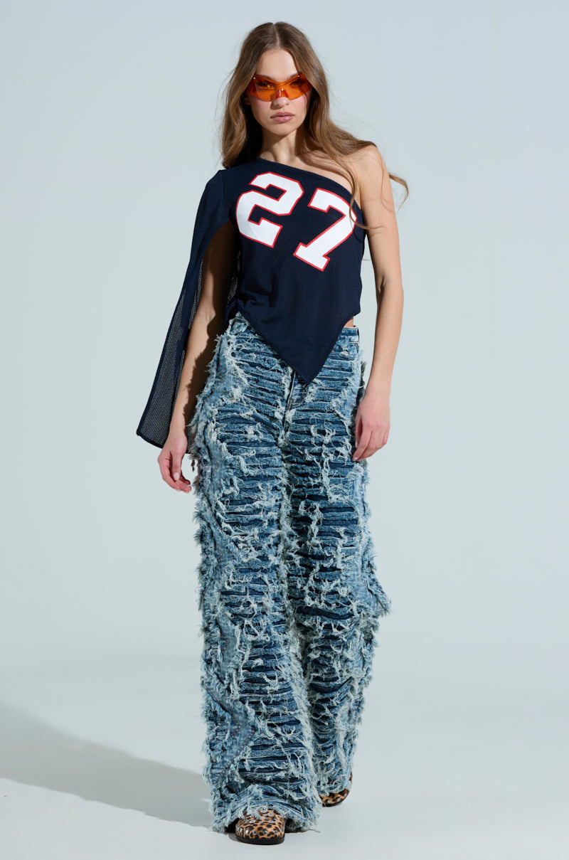 Wearing the GAME TIME JERSEY, sporting an asymmetrical black one-shoulder design with the number 27, paired with textured blue wide-leg jeans showcasing a frayed pattern and vibrant orange sunglasses, they strike a confident pose against a light backdrop. 6