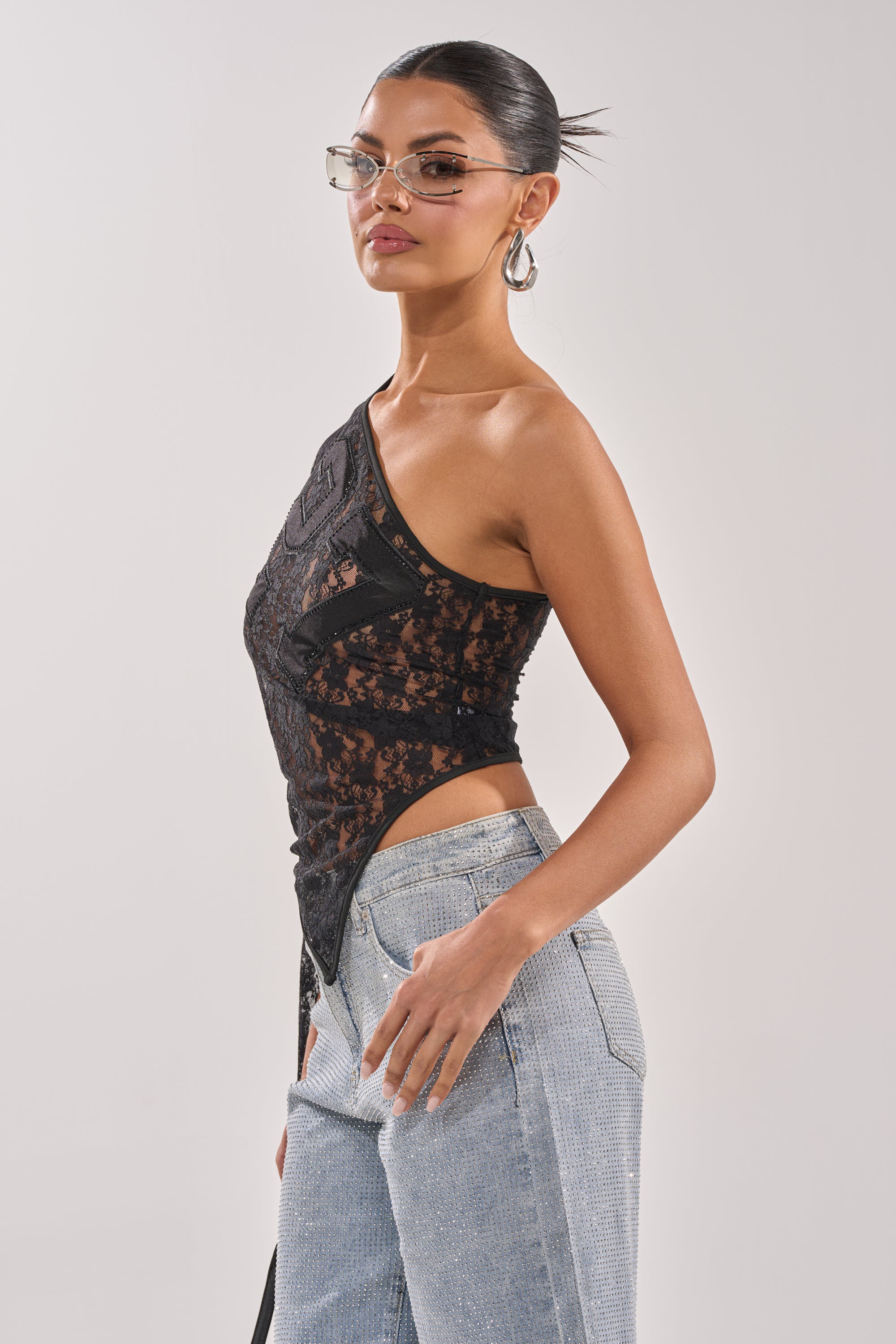 A woman wearing glasses and the GET IT DONE LACE JERSEY, a sheer black lace one-shoulder top, with light blue jeans poses against a light grey background, her hair tied back. 2