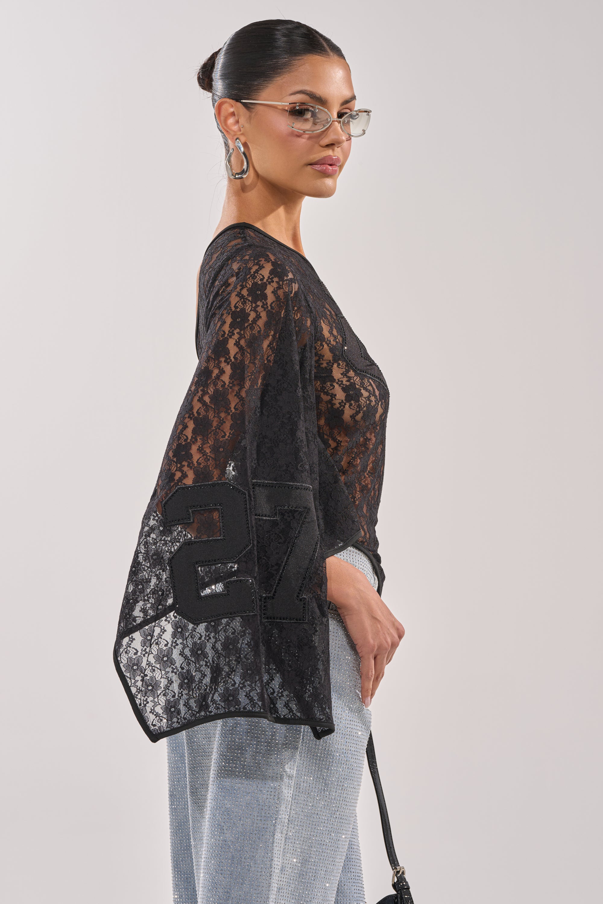 A woman in glasses, hoop earrings, and light blue pants stands in profile, looking over her shoulder. She wears the GET IT DONE LACE JERSEY— a sheer black, one-shoulder lace top with the number 27 on the sleeve—against a light background. 3