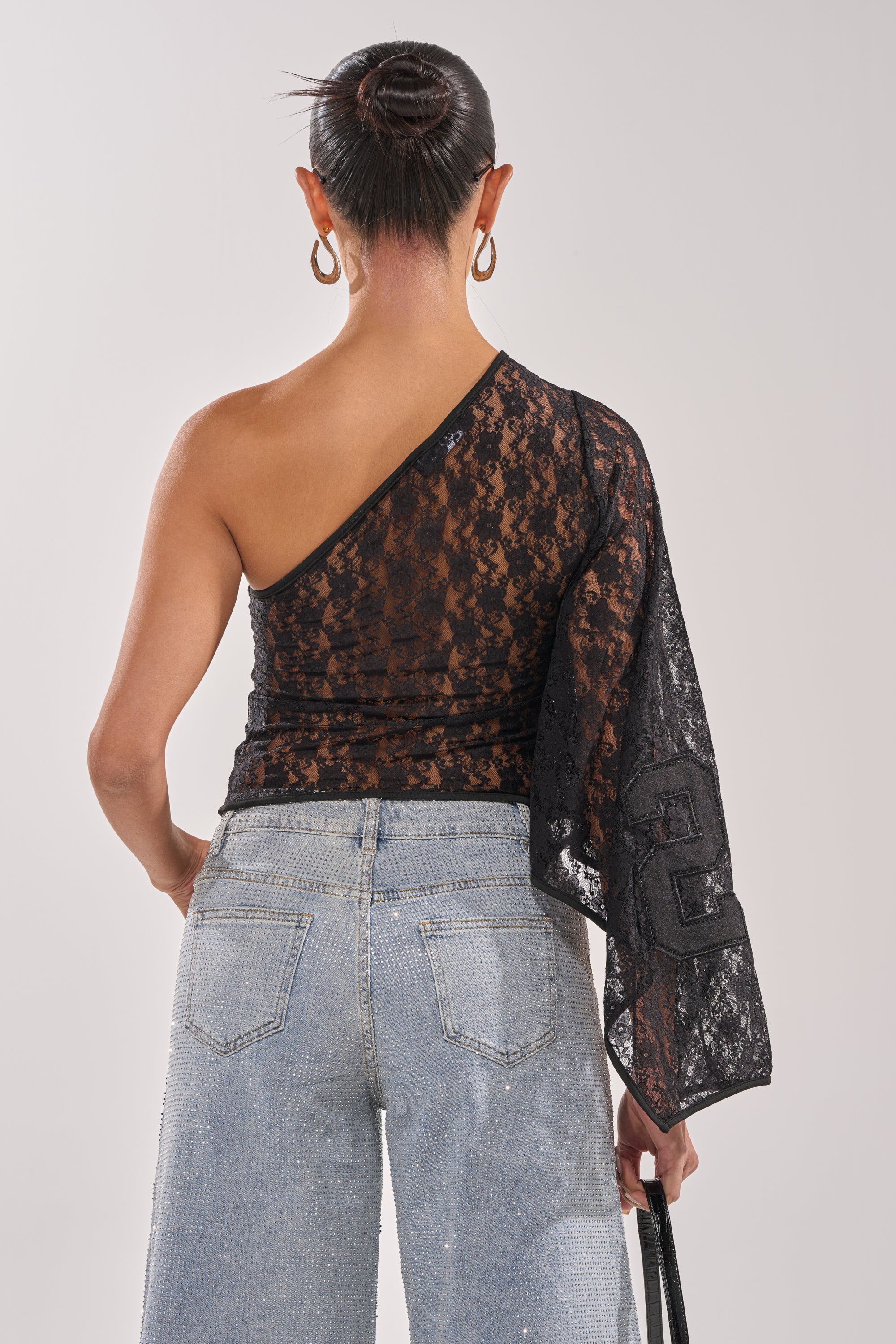 A woman with her hair in a neat bun stands with her back to the camera, wearing light blue jeans and the GET IT DONE LACE JERSEY—a sheer black one-shoulder top with a single long sleeve—while holding a handbag by her side. 4
