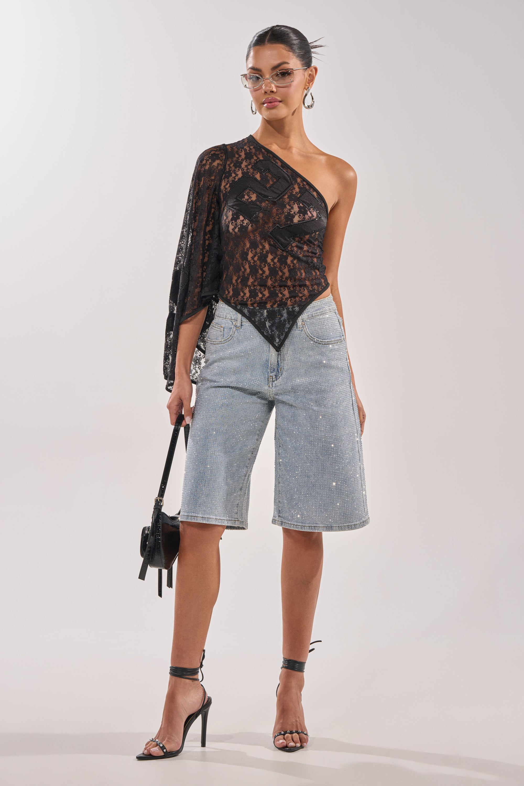 A woman wearing the GET IT DONE LACE JERSEY—a sheer black, one-sleeve lace top—paired with long denim cutoffs, black high-heeled sandals, clear-rimmed glasses, and carrying a small black handbag. 5