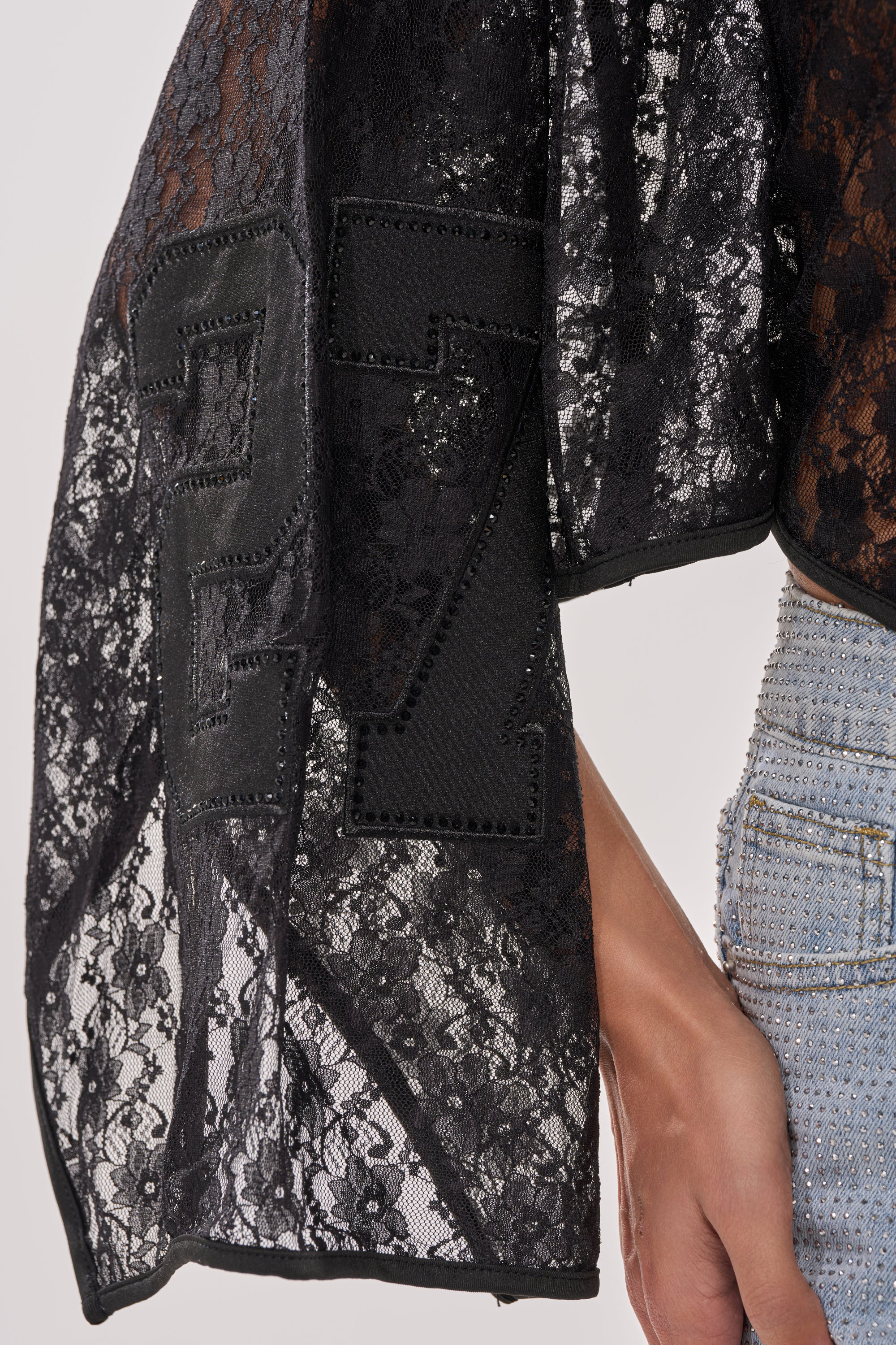 A close-up of a person wearing the GET IT DONE LACE JERSEY—a sheer black lace top with rhinestone accents and "27" on the sleeve—paired with light blue studded jeans. Their hand rests by their side. 7