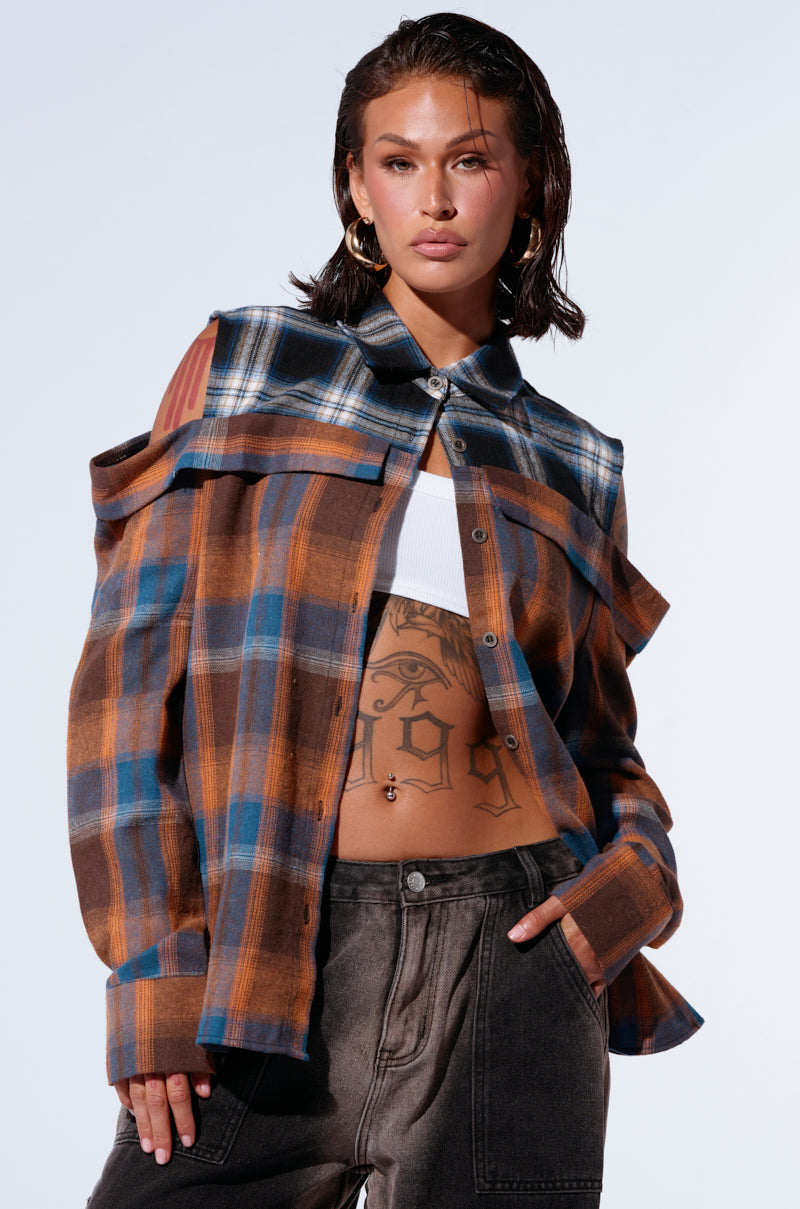 A person with wet-look hair stands confidently, wearing the ALL IN A DAYS WORK BUTTON DOWN FLANNEL SHIRT, a stylish two-tone plaid shirt with cut-out shoulder details over a white crop top. They have a tattoo on their midriff and are posing with one hand in the pocket of their high-waisted, faded jeans. 0