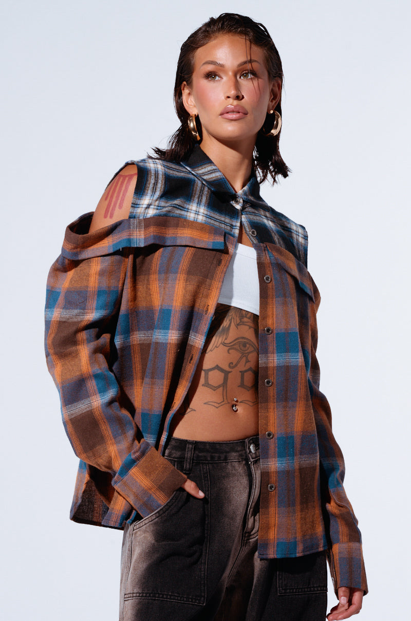 A person with wet, slicked-back hair models the "All in a Days Work Button Down Flannel Shirt," featuring cut-out shoulders and a two-tone plaid design. They wear a white crop top underneath, showcasing several visible tattoos on their chest and stomach. The relaxed silhouette is completed with dark jeans, one hand casually tucked in the pocket. 1