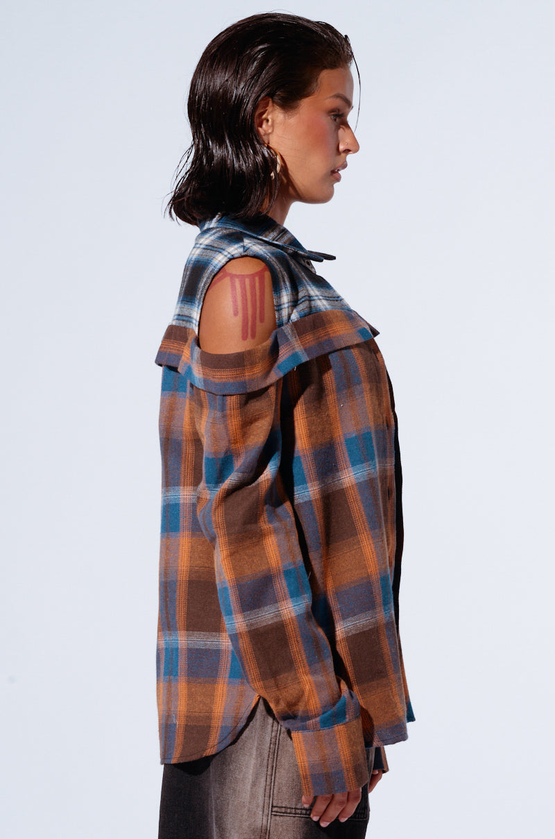 A person with wet, slicked-back hair is standing in profile against a plain background. They are wearing the "ALL IN A DAYS WORK" button-down flannel shirt in blue and brown tones featuring shoulder cutouts, paired with dark-colored pants. A red abstract design is visible on their upper arm, creating a relaxed silhouette. 2