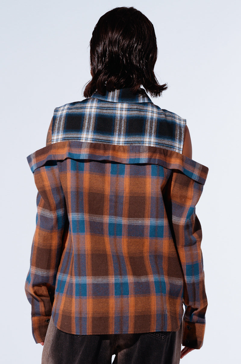 A person with shoulder-length hair faces away from the camera, showcasing a relaxed silhouette of the ALL IN A DAYS WORK BUTTON DOWN FLANNEL SHIRT in shades of brown and blue, paired with dark pants. The background is a plain, light color. 3