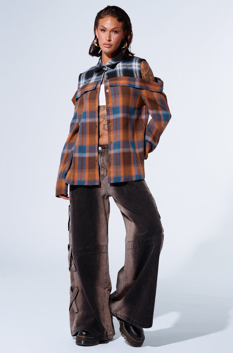 A person with short, wet hair poses confidently against a plain background. They wear the ALL IN A DAYS WORK BUTTON DOWN FLANNEL SHIRT over a revealing top, paired with wide-legged gray pants adorned with side laces. The relaxed silhouette is completed with black shoes. 4