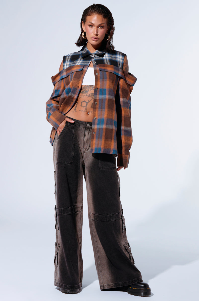 A woman stands confidently against a plain backdrop, showcasing the ALL IN A DAYS WORK BUTTON DOWN FLANNEL SHIRT. This bold two-tone plaid shirt with cut-out shoulders reveals tattoos on her torso. She pairs the button-down flannel with loose, dark cargo pants and black shoes. Her hair is styled back, and she poses with one hand in her pocket. 5