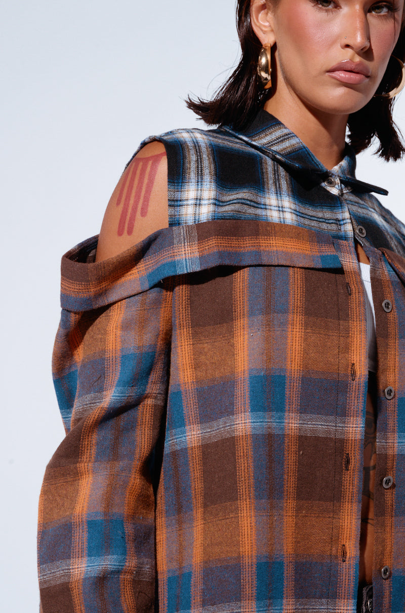 A person poses in the "All In A Day's Work Button Down Flannel Shirt," featuring a two-tone plaid pattern and cut-out shoulders. A unique red stripe tattoo on their left shoulder, a white shirt worn underneath, and gold hoop earrings are visible. The relaxed silhouette and partially shown face highlight contemporary fashion.
 6