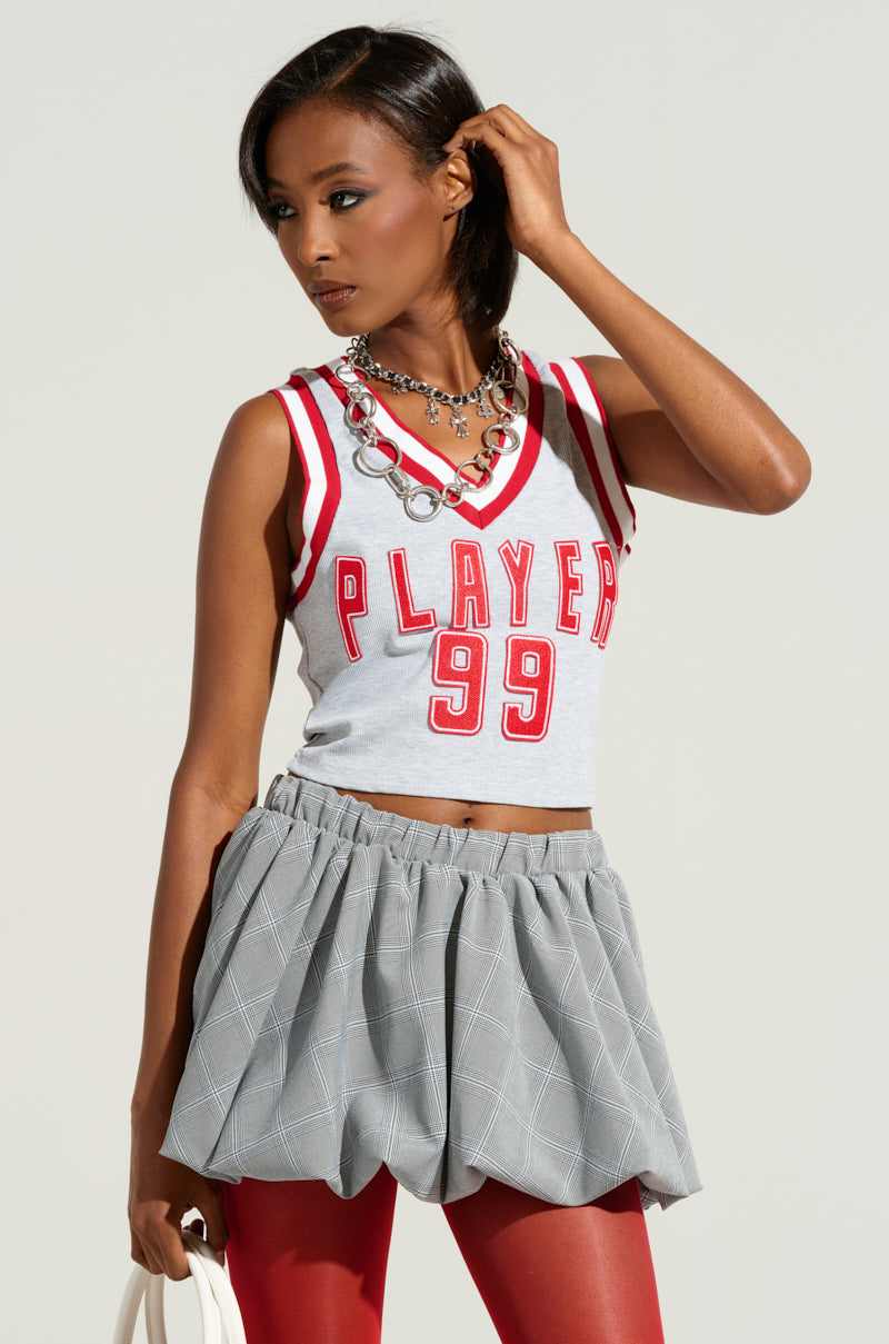 A woman in a sporty chic style ensemble wears the ROSTER GRAPHIC CROPPED TANK featuring "PLAYER 99" against a plain background. She accessorizes with a necklace, plaid skirt, and red leggings while holding a white bag. Her gaze is directed to the side as one hand gently touches her hair. 0