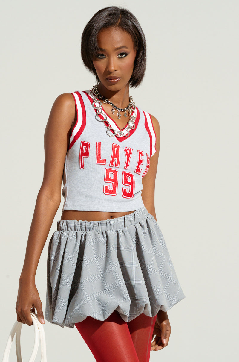 A person exudes a sporty chic style with their ROSTER GRAPHIC CROPPED TANK featuring "Player 99" in red, layered necklaces, a gray plaid bubble skirt, and red leggings. They hold a white bag against a neutral background. 1