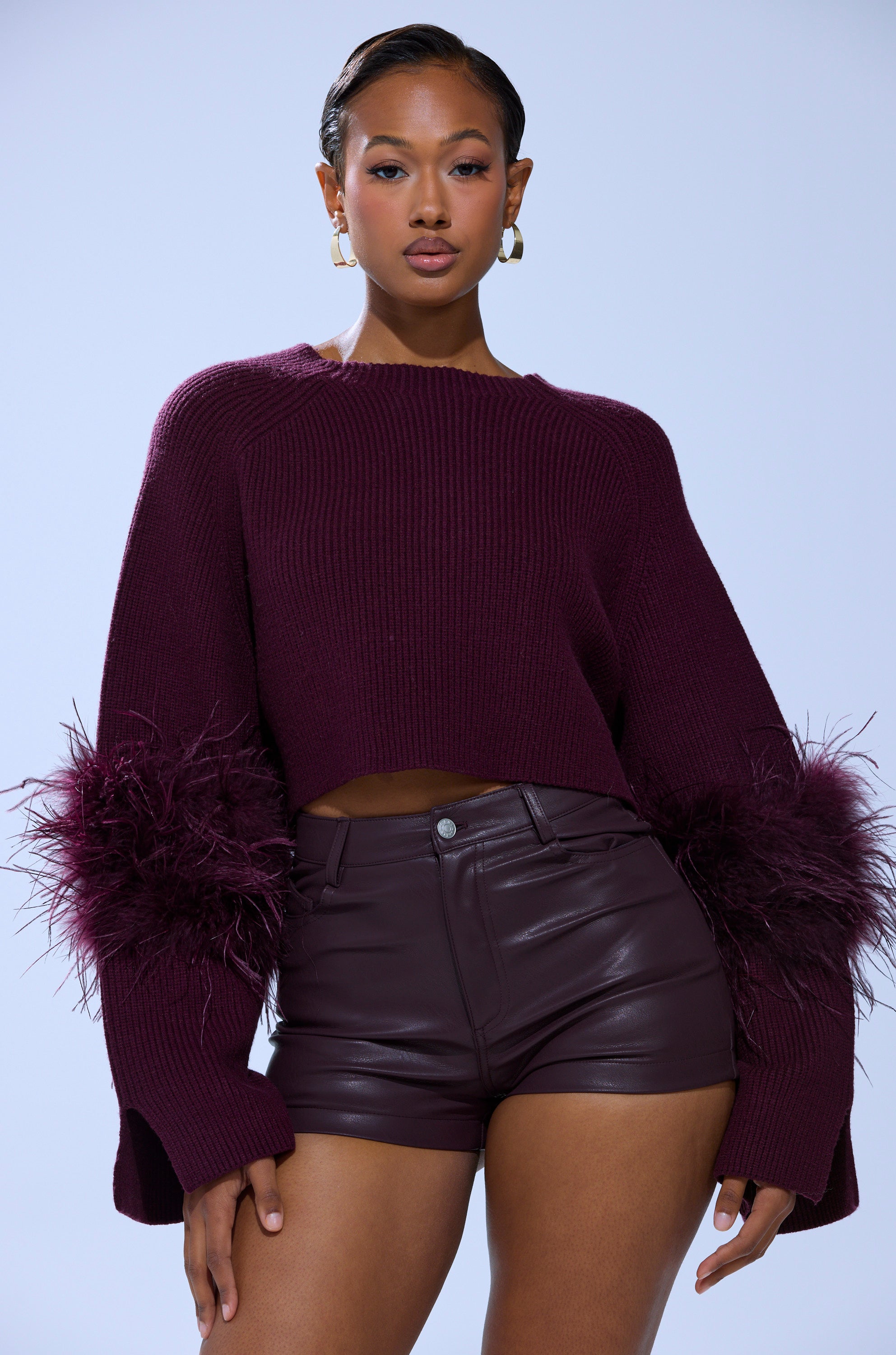 A woman stands confidently against a light background, wearing the MILLY FEATHER TRIM SWEATER with feathered cuffs, gold hoop earrings, and high-waisted leather shorts.