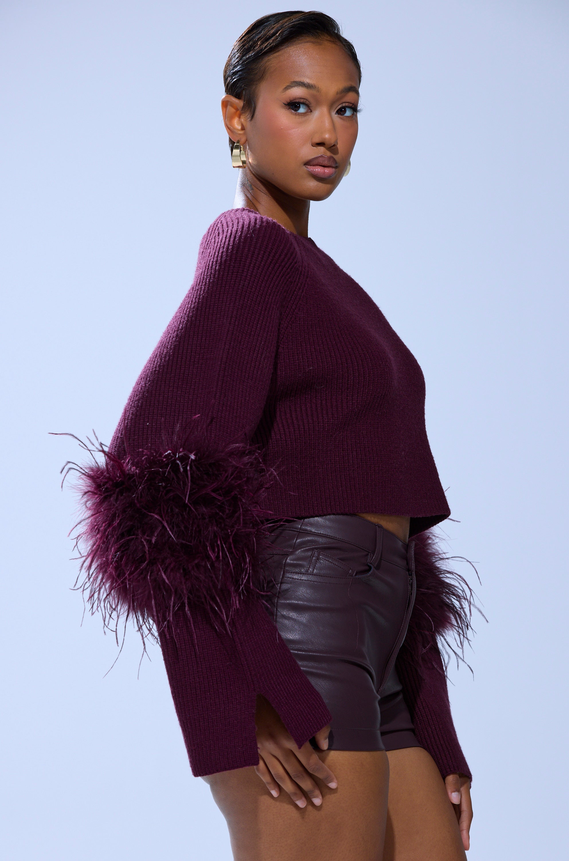 A woman models the MILLY FEATHER TRIM SWEATER, featuring cozy knit fabric and textured burgundy sleeves, paired with high-waisted leather shorts against a light gray background.