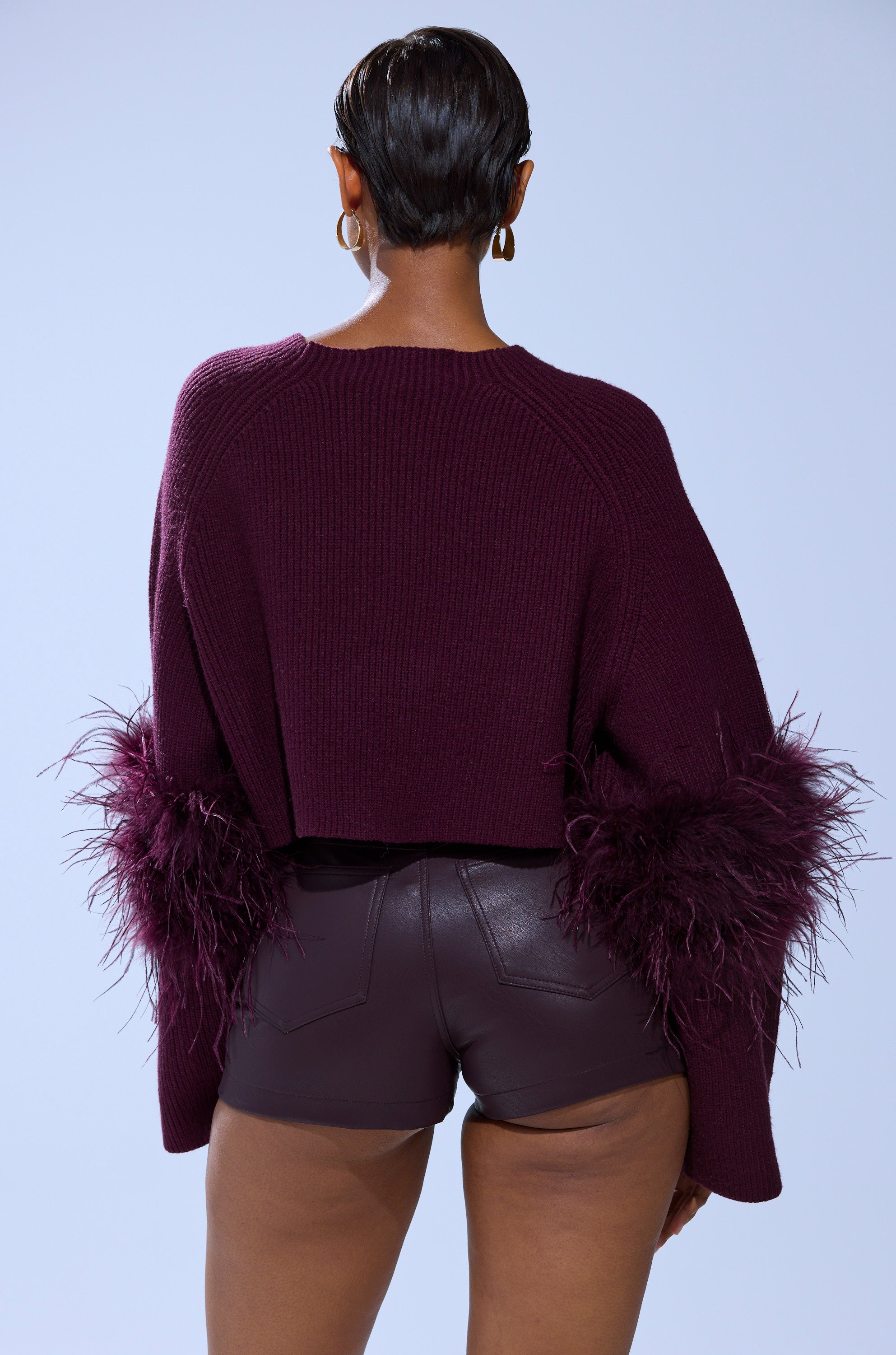 Seen from the back, a person with short hair wears the MILLY FEATHER TRIM SWEATER featuring cozy knit fabric and feathered sleeve accents, paired with high-waisted, dark brown leather shorts.