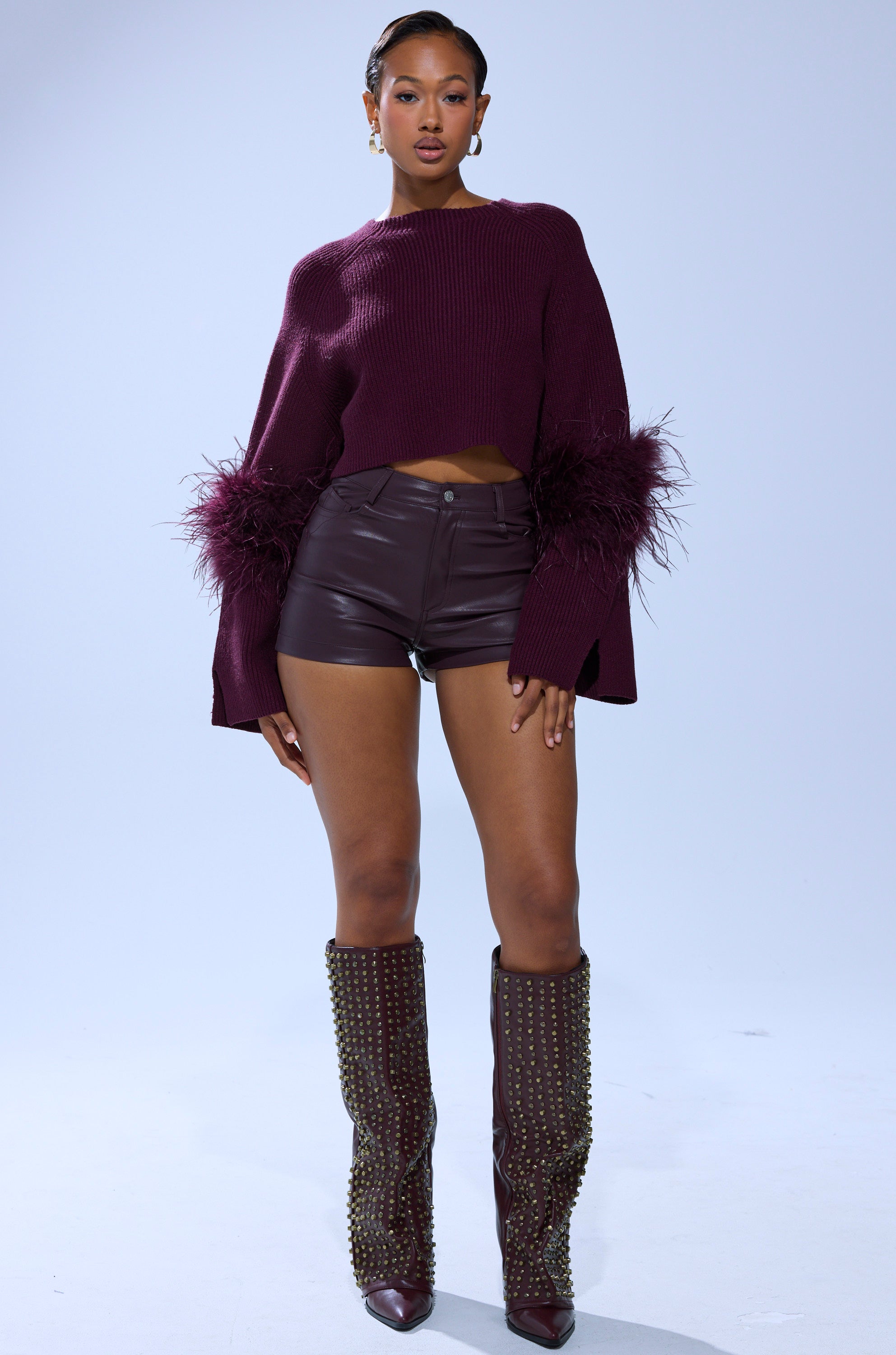 A woman stands confidently in the MILLY FEATHER TRIM SWEATER with burgundy accents, paired with high-waisted leather shorts and gold-studded knee-high boots, set against a plain light background.