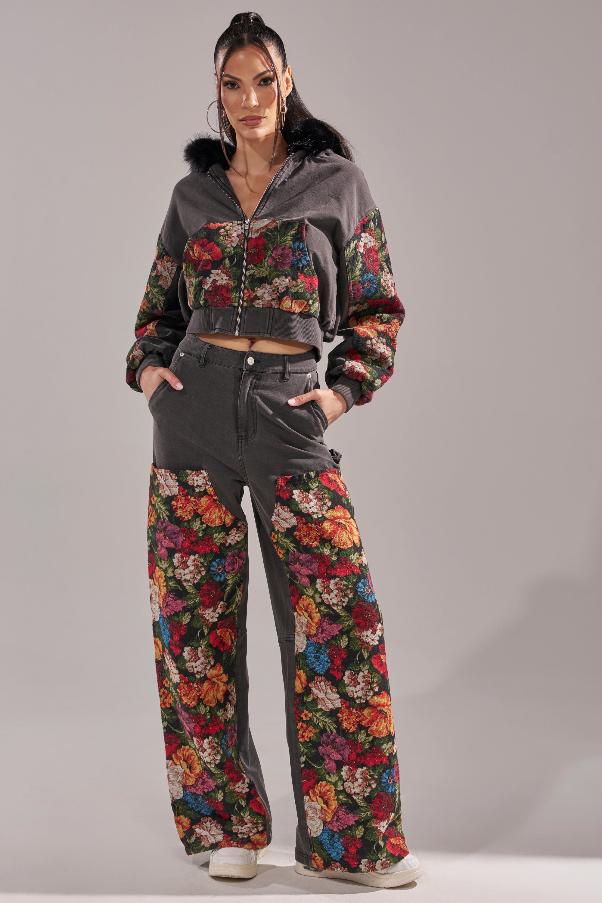 A woman models the TRY ME FLORAL DENIM set—a gray, floral print tracksuit with a cropped zip-up hoodie and wide-leg pants—paired with hoop earrings and white sneakers against a light gray background. 0