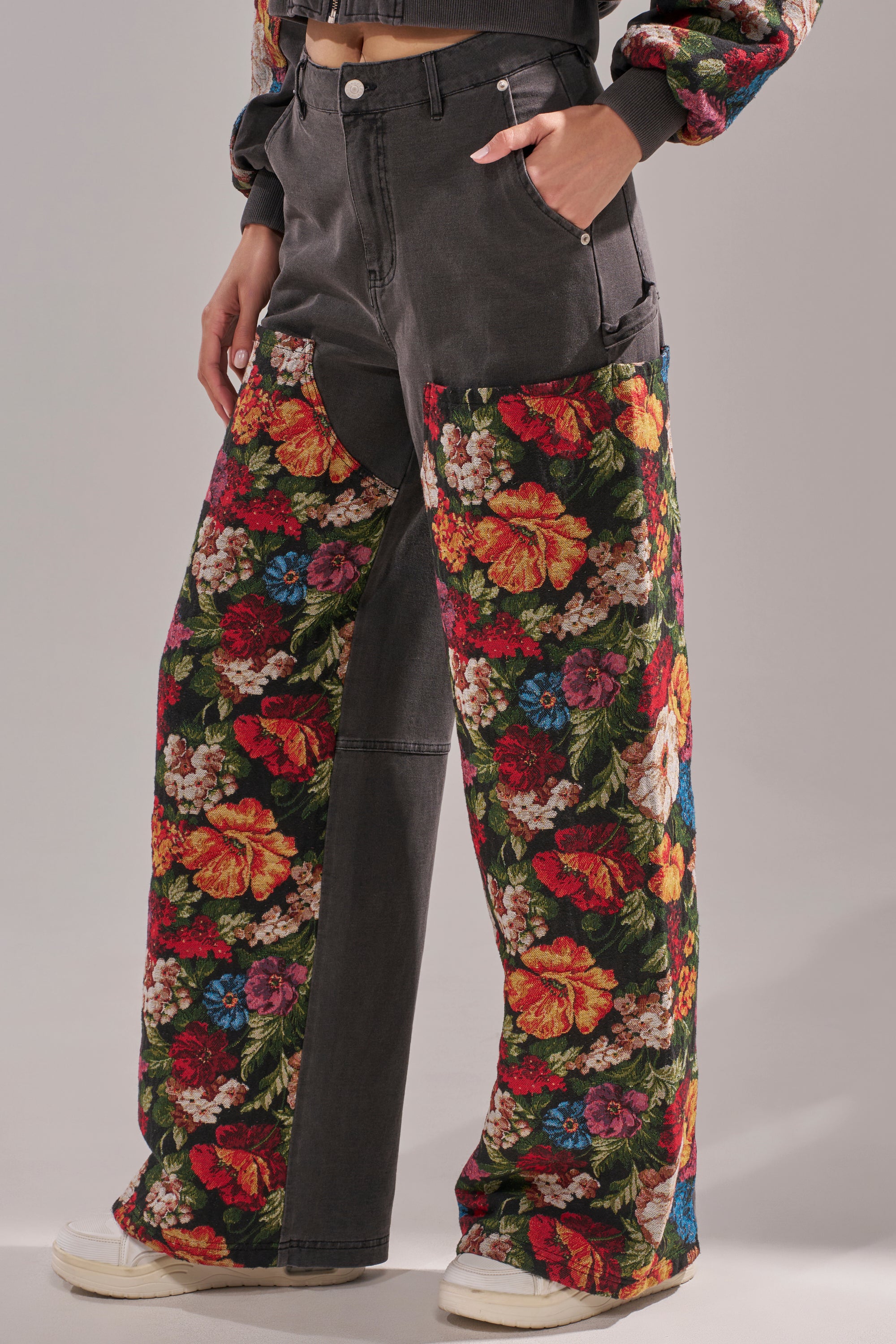 Model wears the TRY ME FLORAL DENIM—high-waisted, wide-leg jeans with vibrant multicolored floral panels on the lower half. White sneakers complete the look, making these statement jeans stand out against a neutral backdrop. 2