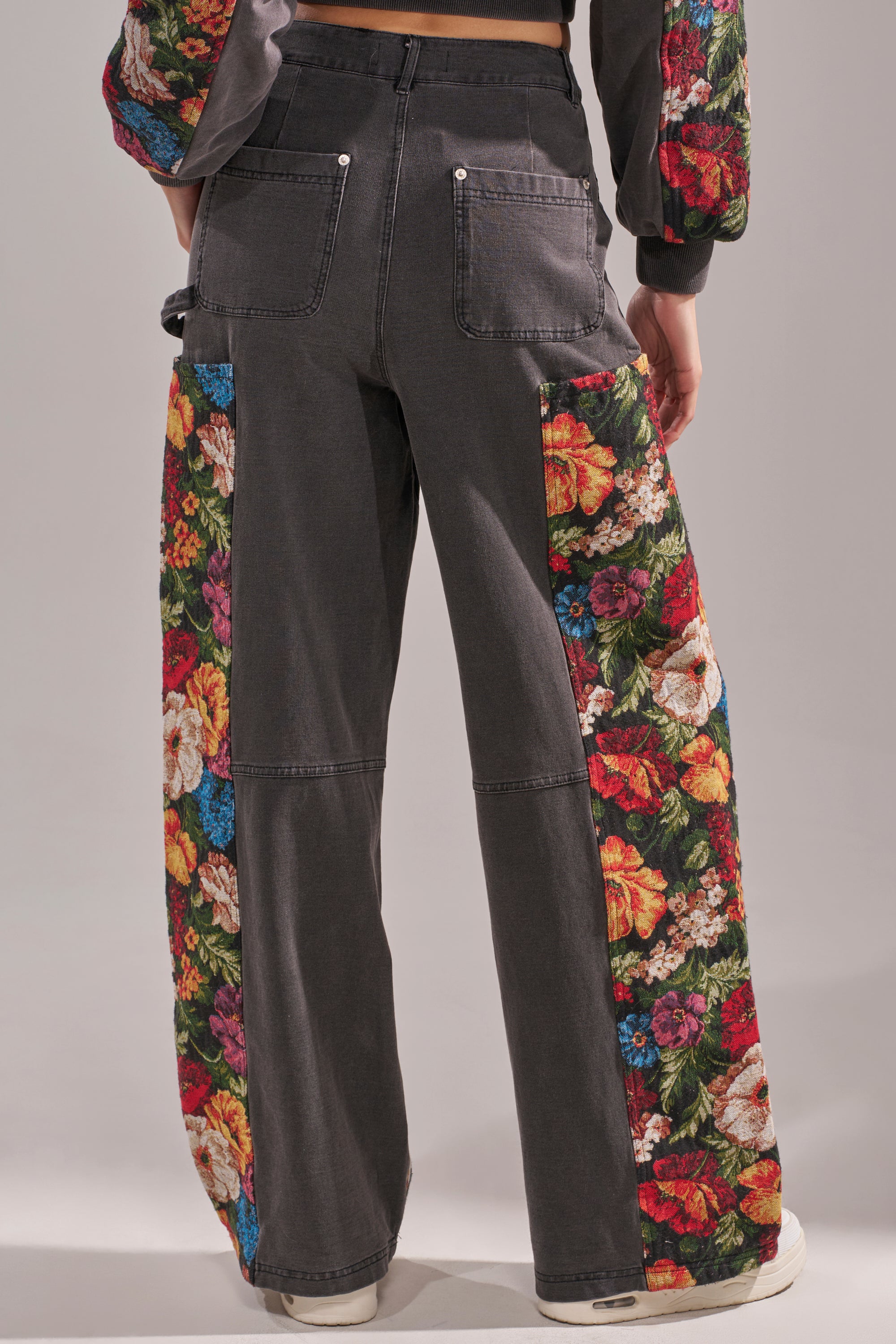 Someone wearing the TRY ME FLORAL DENIM, featuring high-waisted jeans with colorful floral fabric panels on the sides and back pockets, paired with white sneakers. Photo captured from behind. 3
