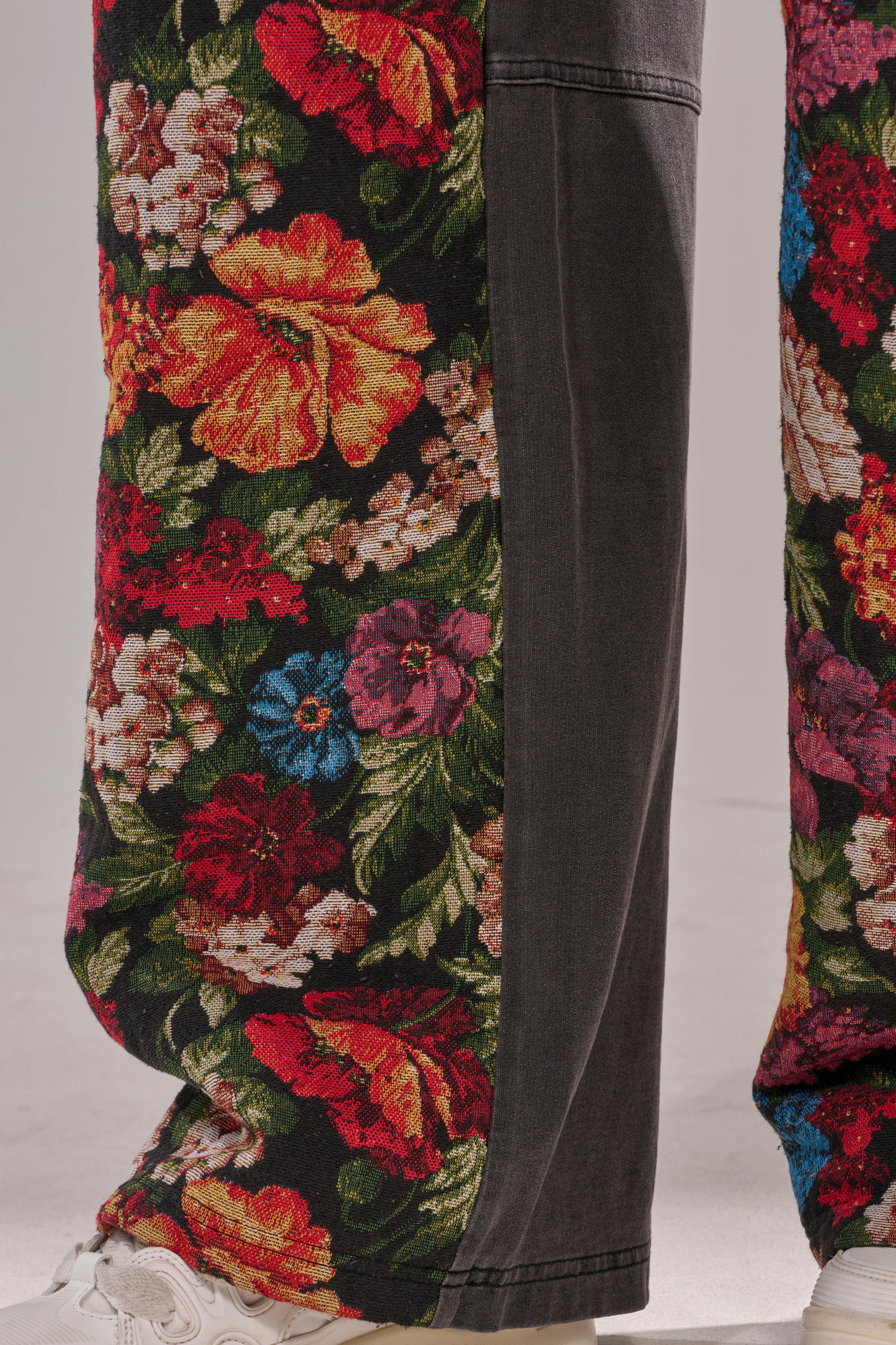 A close-up of the TRY ME FLORAL DENIM high-waisted jeans with a vibrant floral tapestry in red, yellow, and blue, contrasted by black denim panels and styled with white sneakers. 7