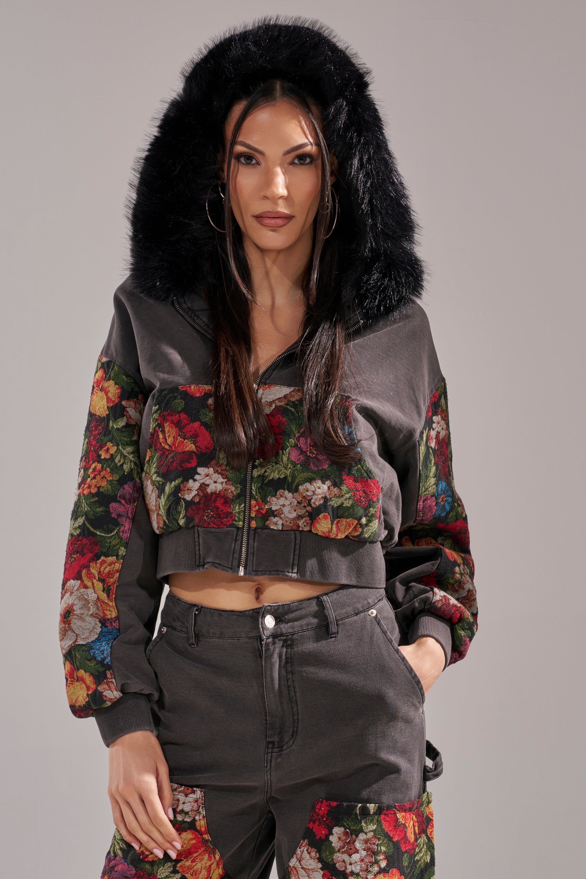 A woman wearing the TRY ME FLORAL DENIM ZIP UP SWEATSHIRT with a black faux fur-trimmed hood and colorful floral patterns, paired with matching high-waisted pants, stands against a plain light background. 0