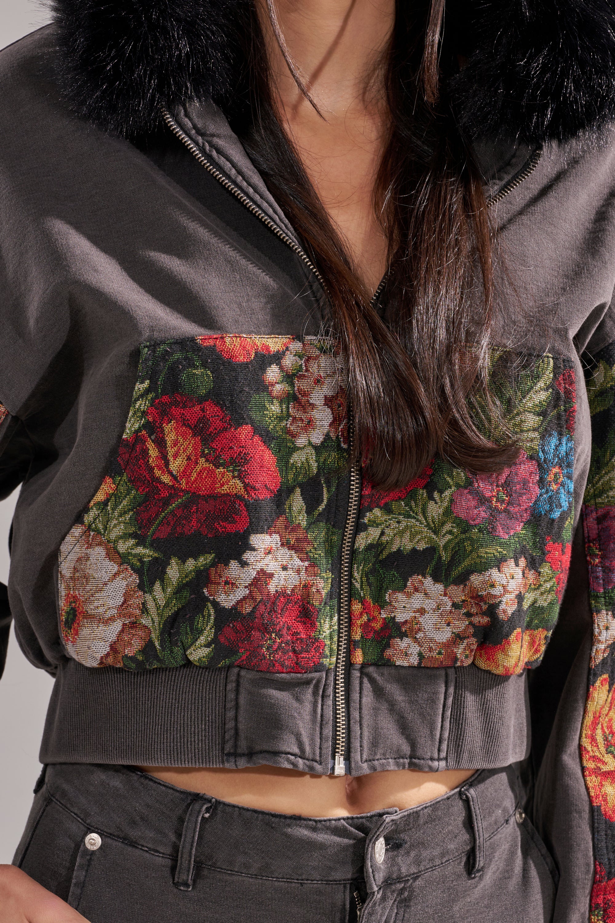 A woman with long dark hair and pants wears the TRY ME FLORAL DENIM ZIP UP SWEATSHIRT, featuring a cropped fit, colorful floral denim chest pocket, and black faux fur collar trim. Only her torso is visible. 10