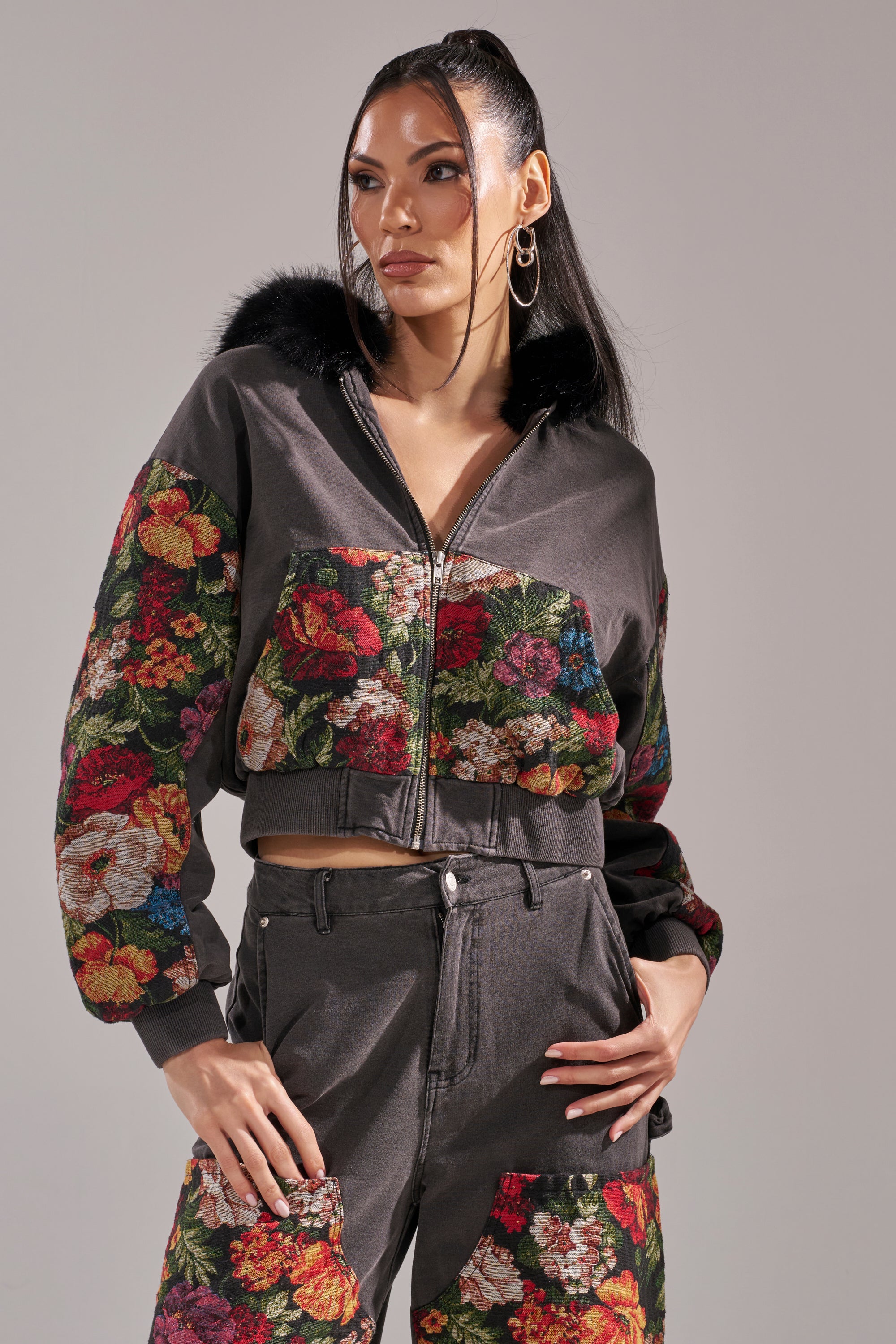 A woman with long dark hair in a high ponytail wears the TRY ME FLORAL DENIM ZIP UP SWEATSHIRT, featuring vibrant red and pink patterns. She accessorizes with hoop earrings and stands with one hand on her hip, looking to the side. 1