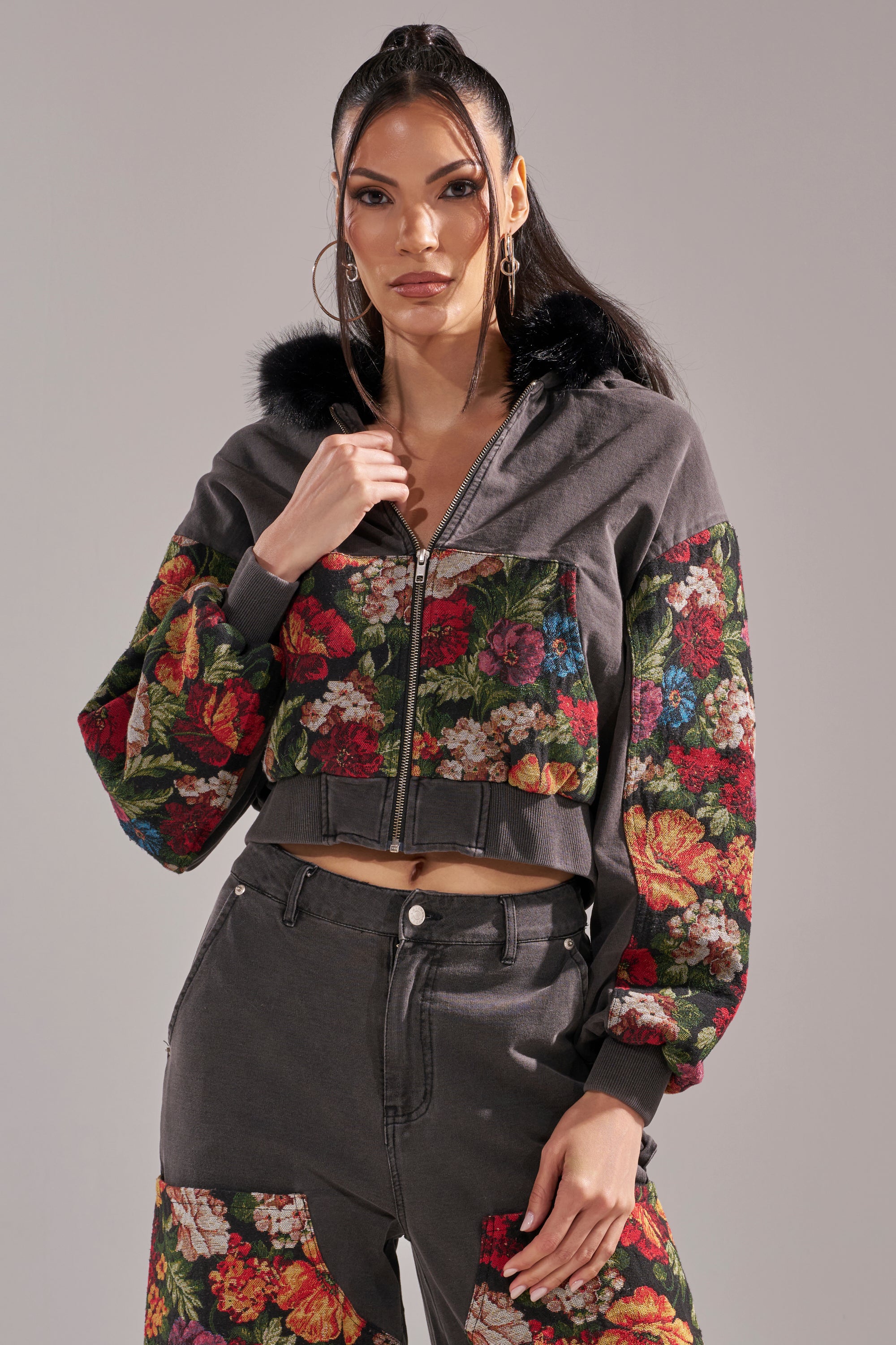 A woman stands confidently in the TRY ME FLORAL DENIM ZIP UP SWEATSHIRT, featuring a dark cropped silhouette with black faux fur hood and vibrant floral-patterned sleeves, paired with matching pants with floral patches. Her long dark hair is in a high ponytail. 2