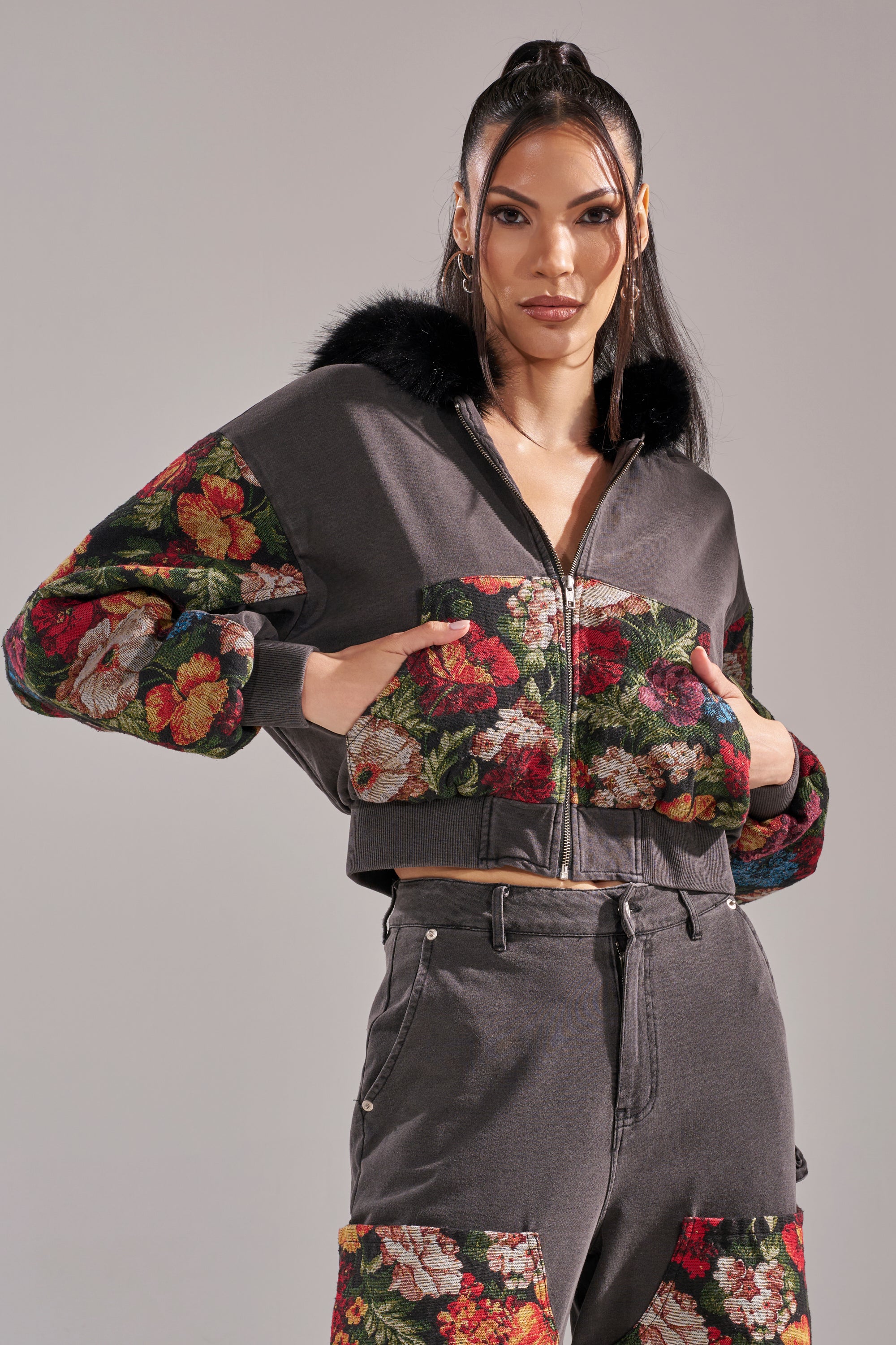 A woman stands confidently with her hands on her TRY ME FLORAL DENIM ZIP UP SWEATSHIRT, a stylish gray cropped hoodie featuring floral-patterned sleeves, pockets, and pant patches, looking directly at the camera. 3