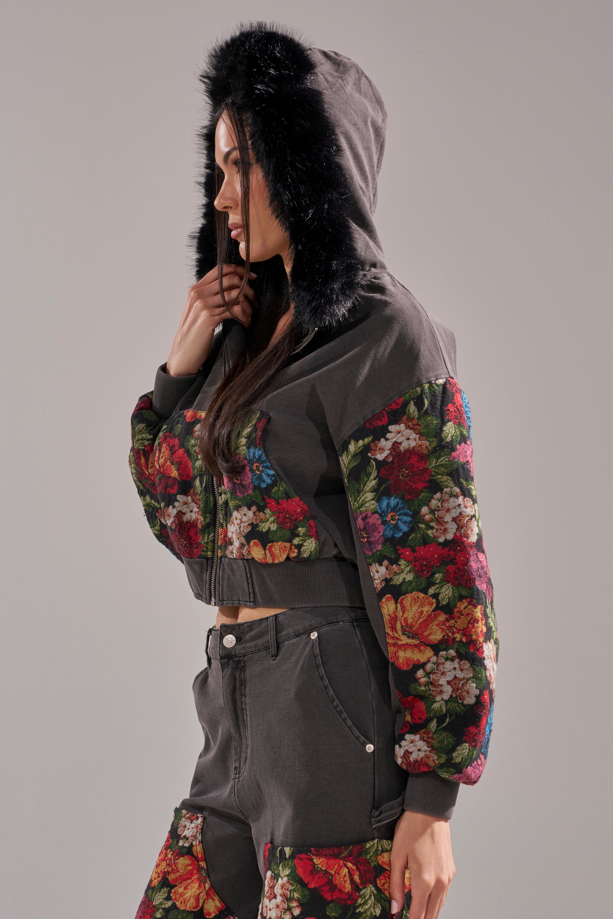A woman stands in profile against a plain background wearing the TRY ME FLORAL DENIM ZIP UP SWEATSHIRT, a dark cropped sweatshirt with a faux fur hood and floral-patterned sleeves, paired with matching floral-accented high-waisted shorts. 5
