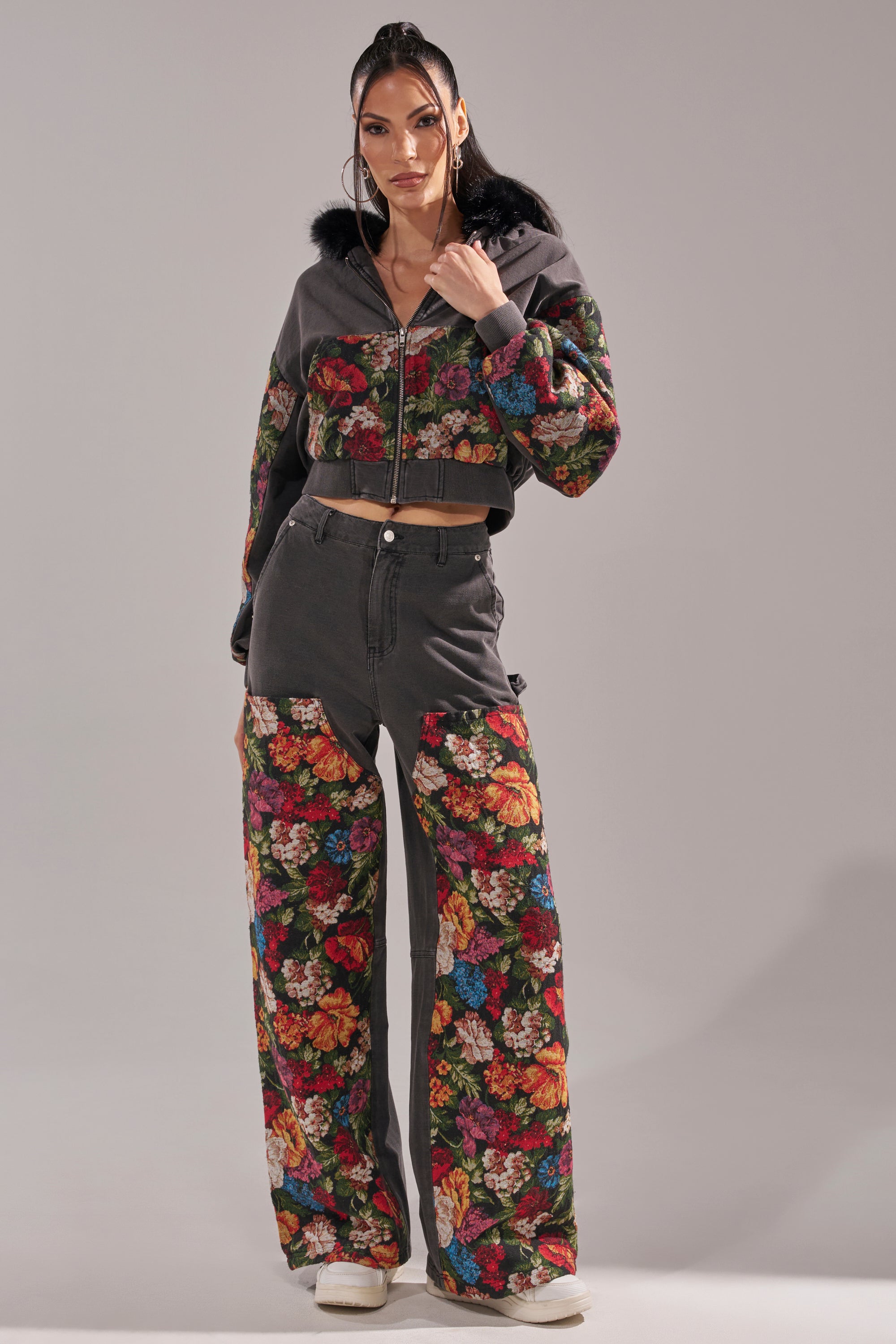 A woman poses in the TRY ME FLORAL DENIM ZIP UP SWEATSHIRT, a black two-piece with vibrant floral prints and a cropped faux fur hooded jacket, paired with loose pants and white sneakers. 8