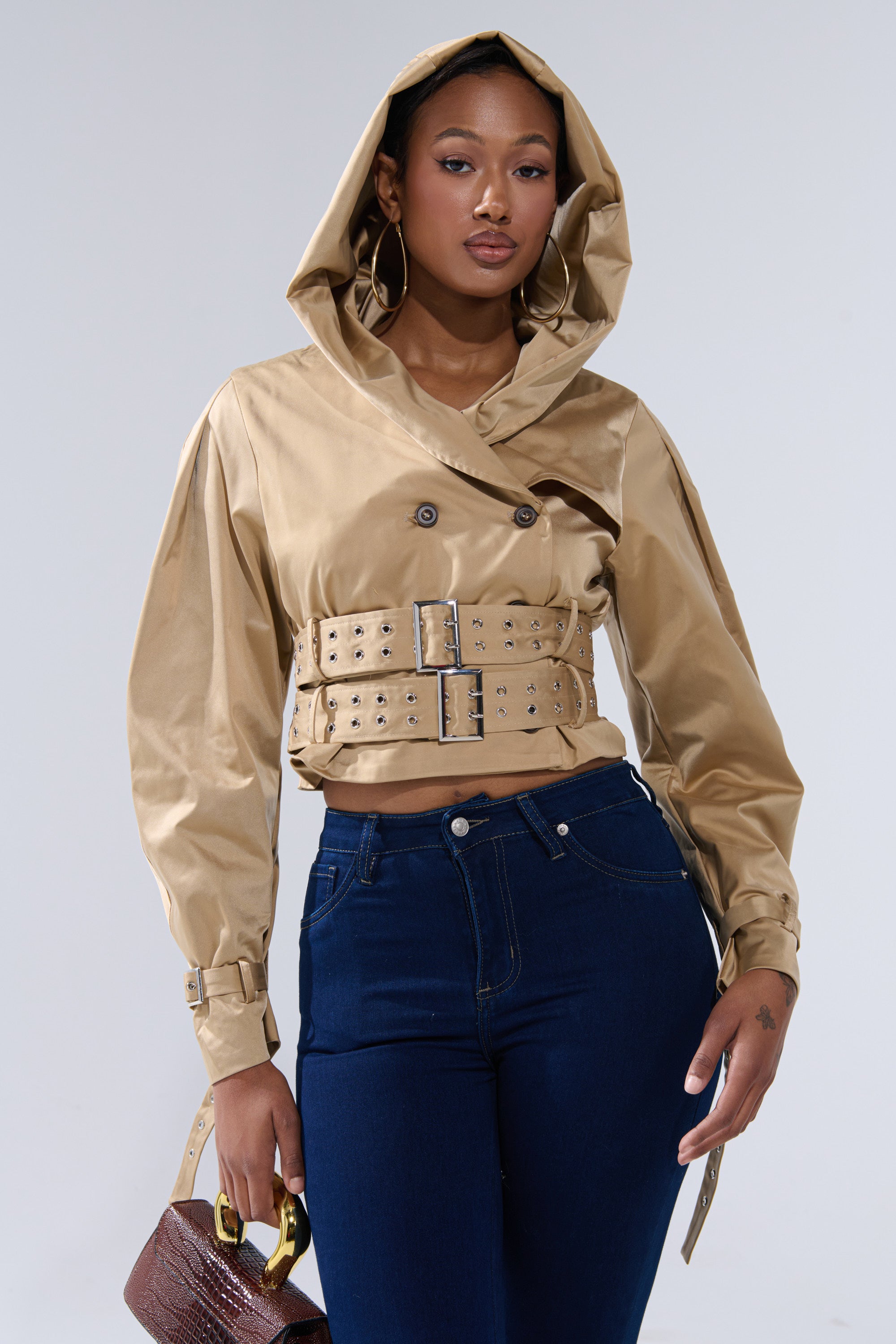 A woman models the ON DUTY CROPPED JACKET, a tan belted piece with an oversized hood, styled with dark blue high-waisted jeans and a small brown textured handbag. She wears hoop earrings and stands against a plain background. 0