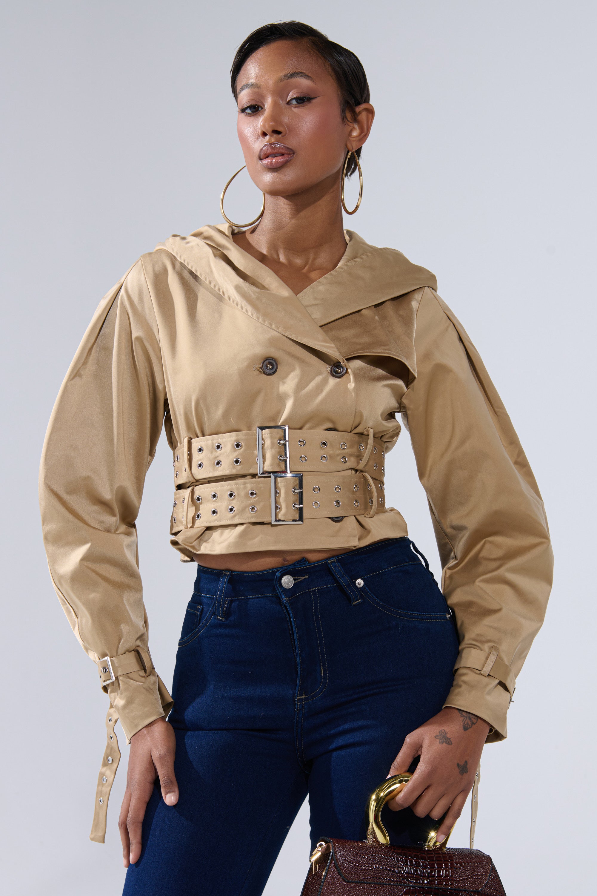 A stylish person poses confidently in the ON DUTY CROPPED JACKET, featuring a tan belted trench silhouette with metallic details, paired with dark blue high-waisted jeans, large hoop earrings, and a textured brown handbag against a plain background. 1