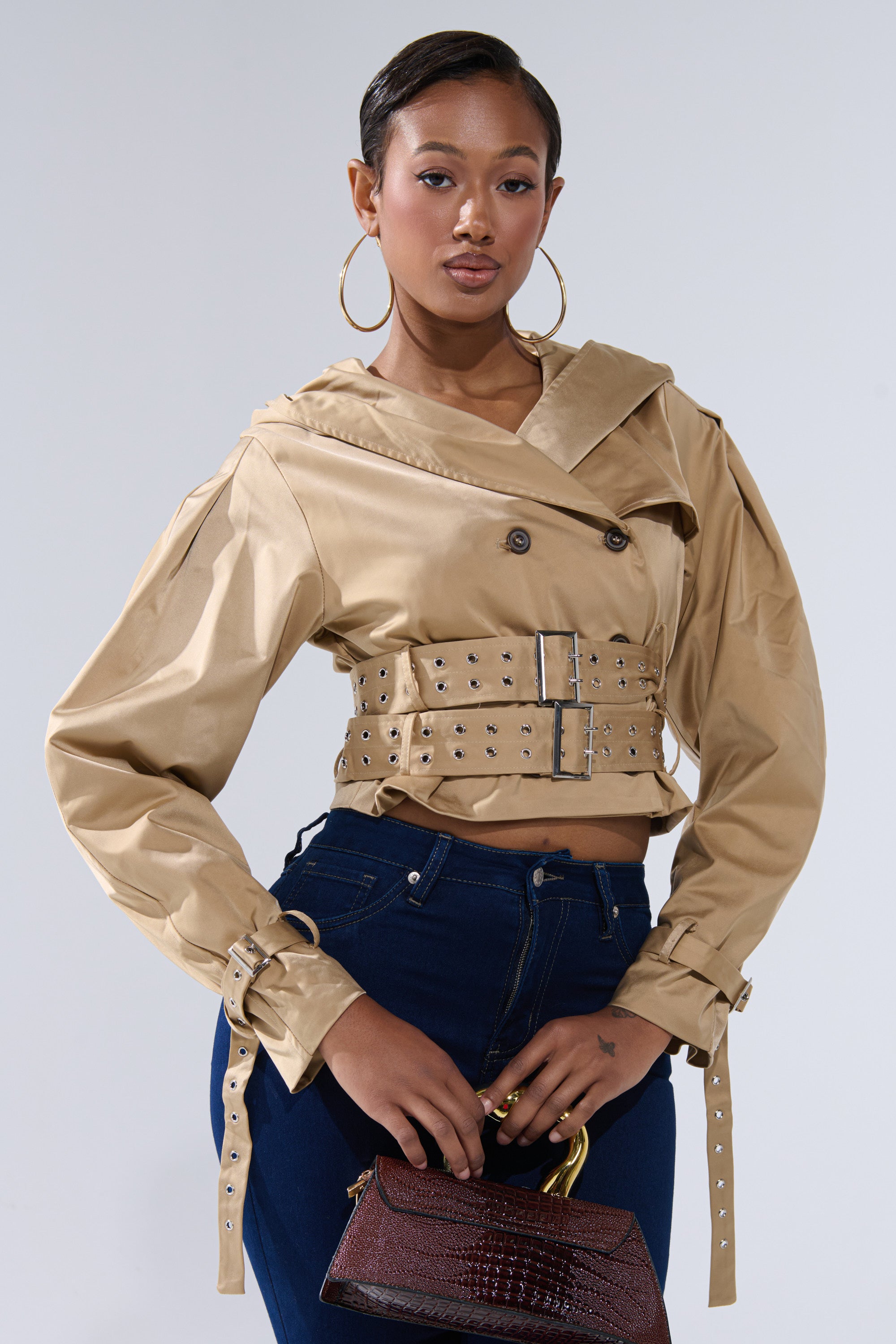 A stylish woman poses against a plain background wearing the ON DUTY CROPPED JACKET—a beige belted jacket with stud details and a trench silhouette—paired with dark blue high-waisted jeans, hoop earrings, and a small brown handbag. 2
