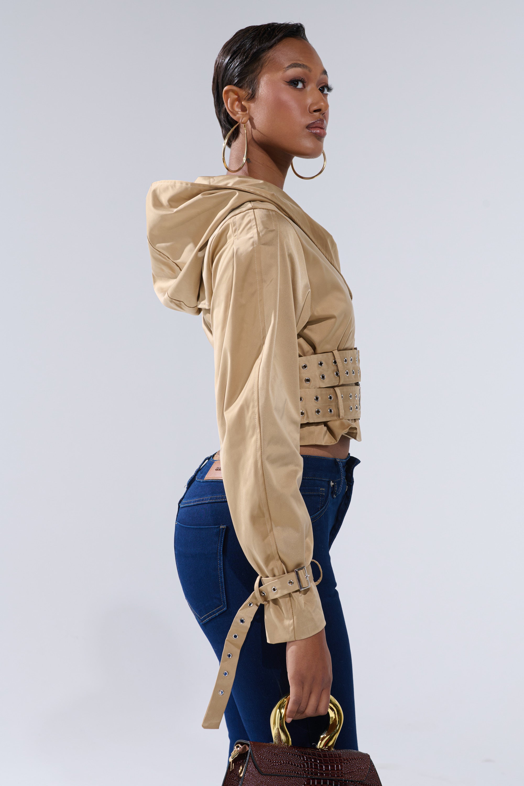A woman models the ON DUTY CROPPED JACKET in tan with belted cuffs, paired with dark blue jeans and large hoop earrings, holding a brown handbag with gold handles against a plain light background. 3