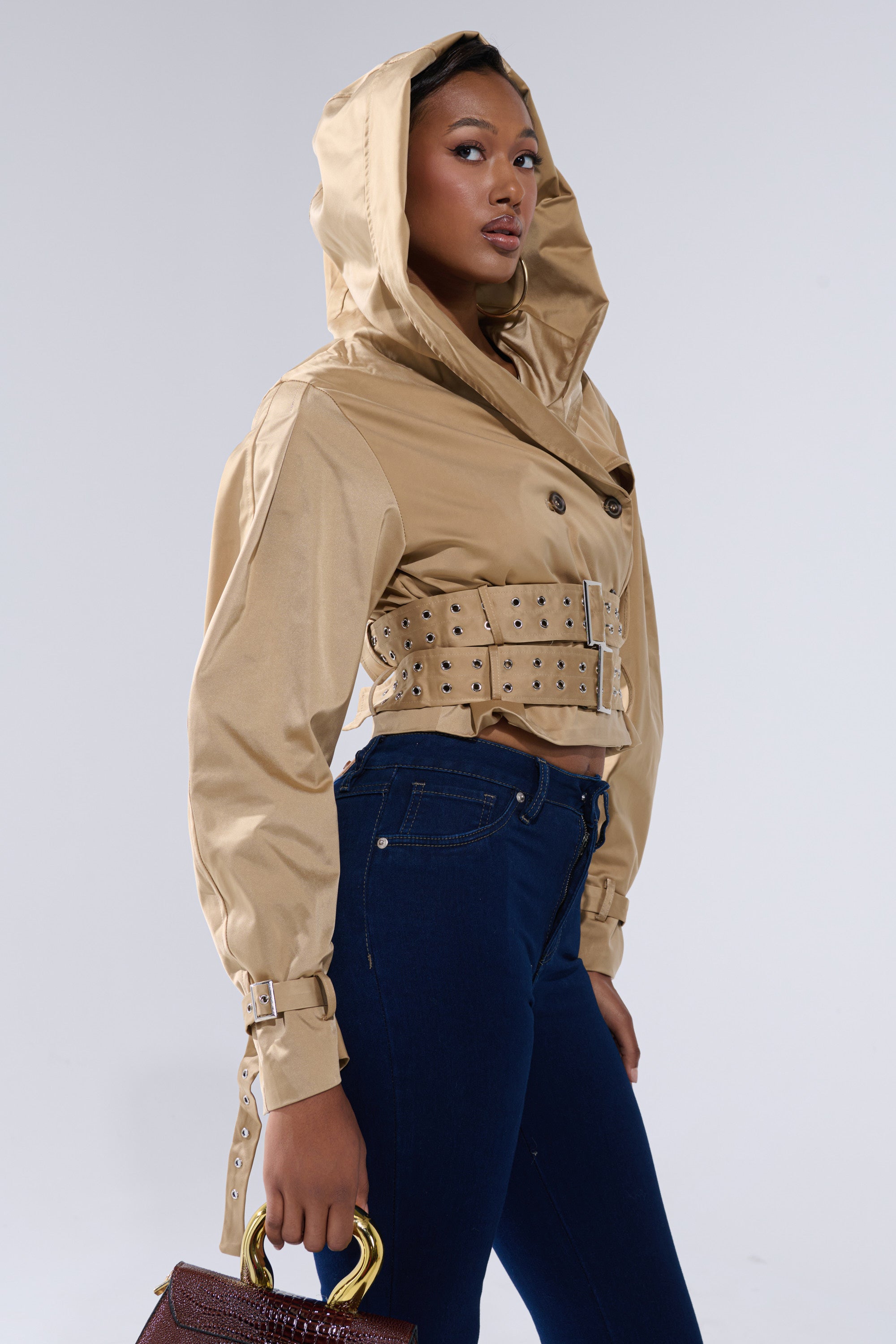 A woman stands against a plain light gray background wearing the ON DUTY CROPPED JACKET with an oversized hood, studded belts, dark blue high-waisted jeans, and holding a brown handbag. 4