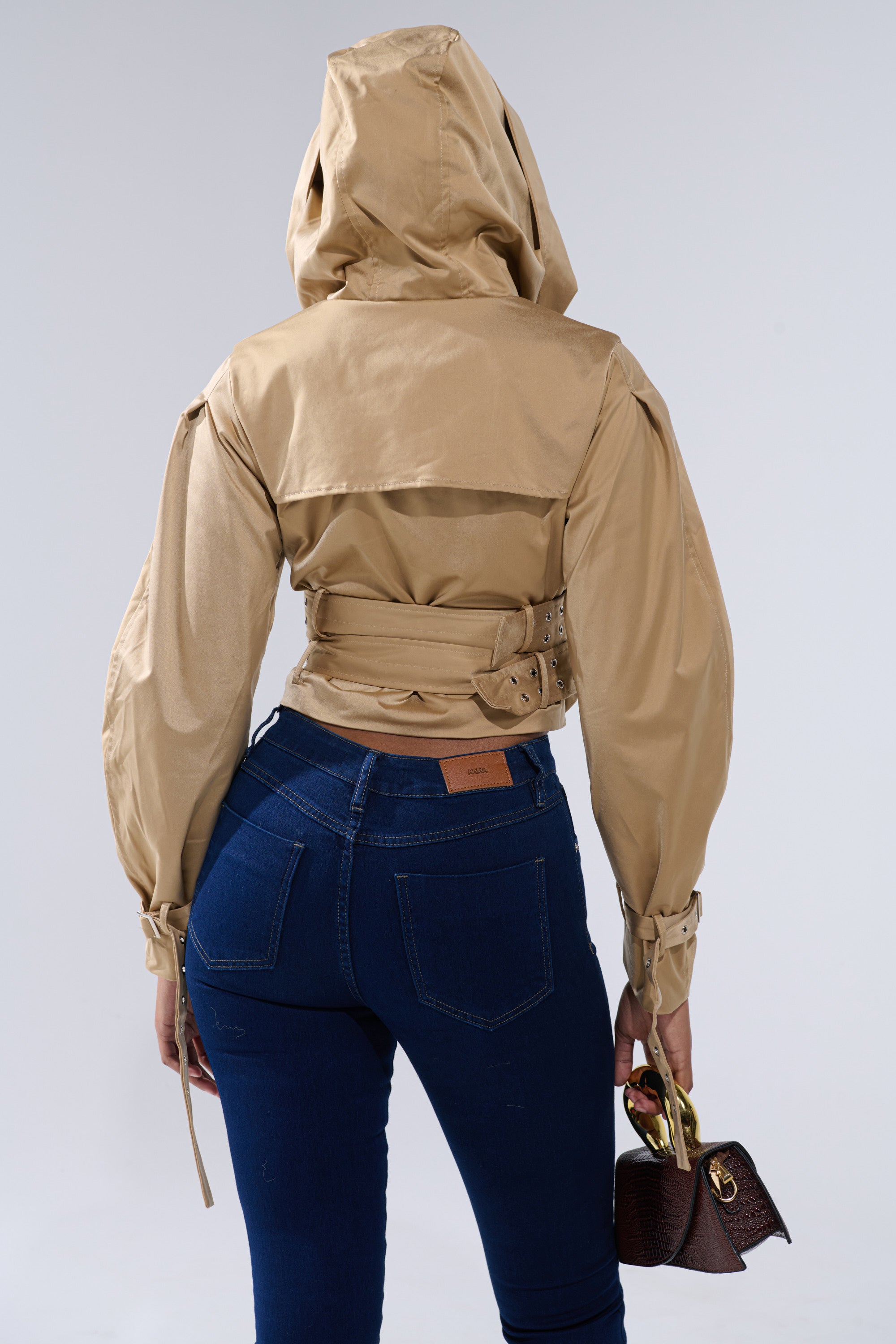 A person stands facing away, wearing the ON DUTY CROPPED JACKET—tan, cropped with an oversized hood and belted waist—paired with blue jeans and holding a small brown handbag. The background is plain light gray. 5