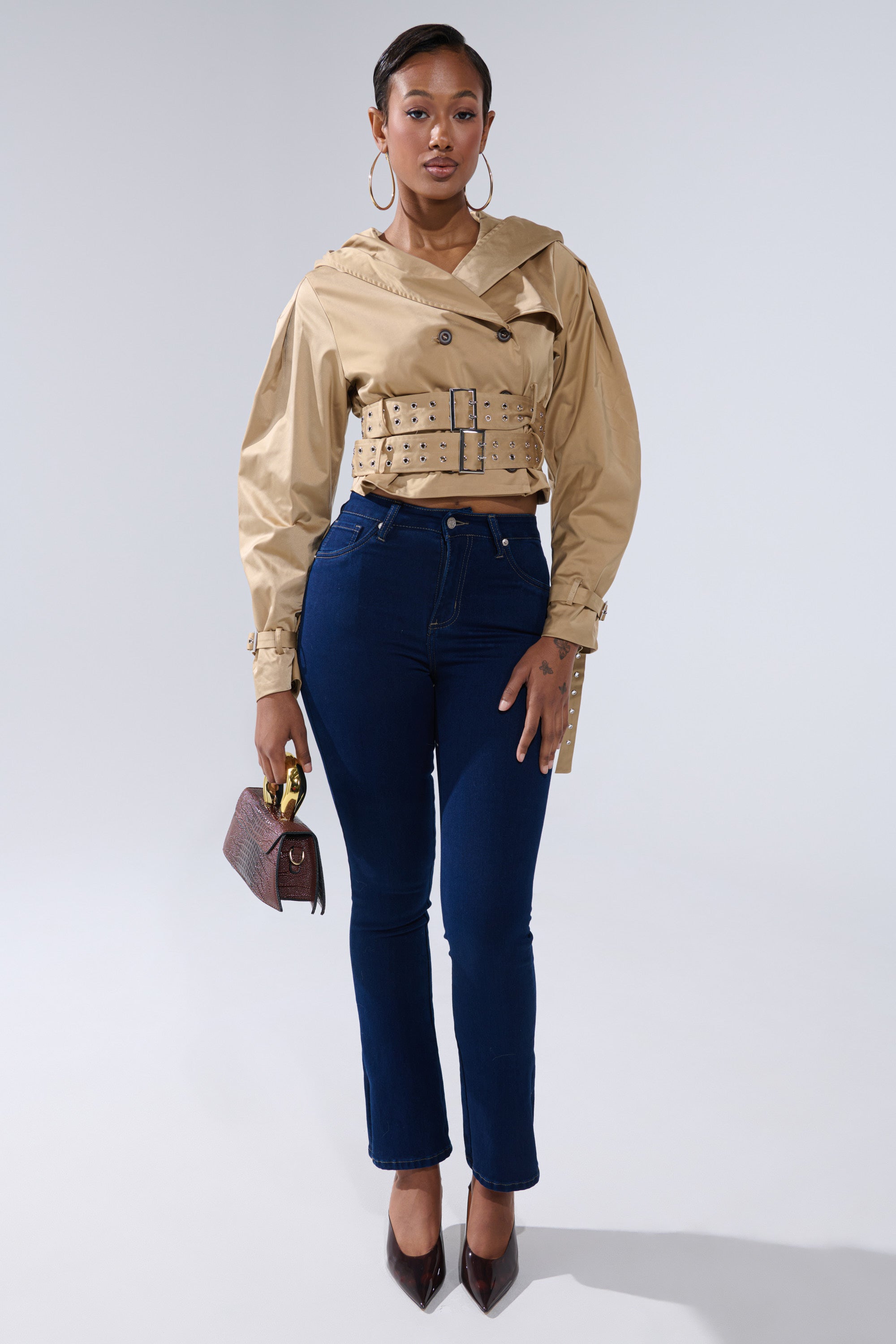 A woman faces forward in the ON DUTY CROPPED JACKET—a tan, belted trench-style piece—paired with dark blue skinny jeans, brown pointed heels, a small brown handbag, and large hoop earrings against a plain light background. 6
