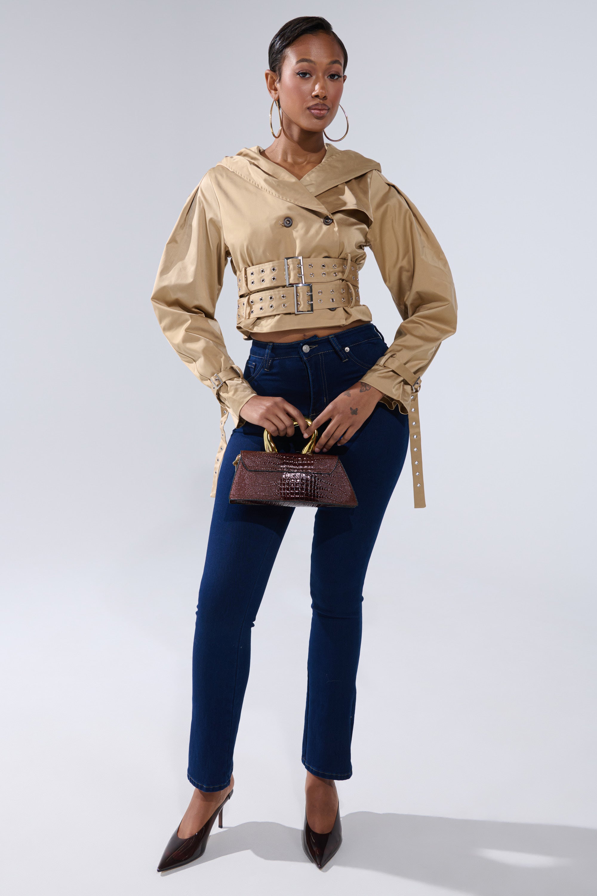 A woman stands confidently in the ON DUTY CROPPED JACKET, styled with dark blue high-waisted jeans, brown pointed heels, a small brown handbag, and large hoop earrings. 7