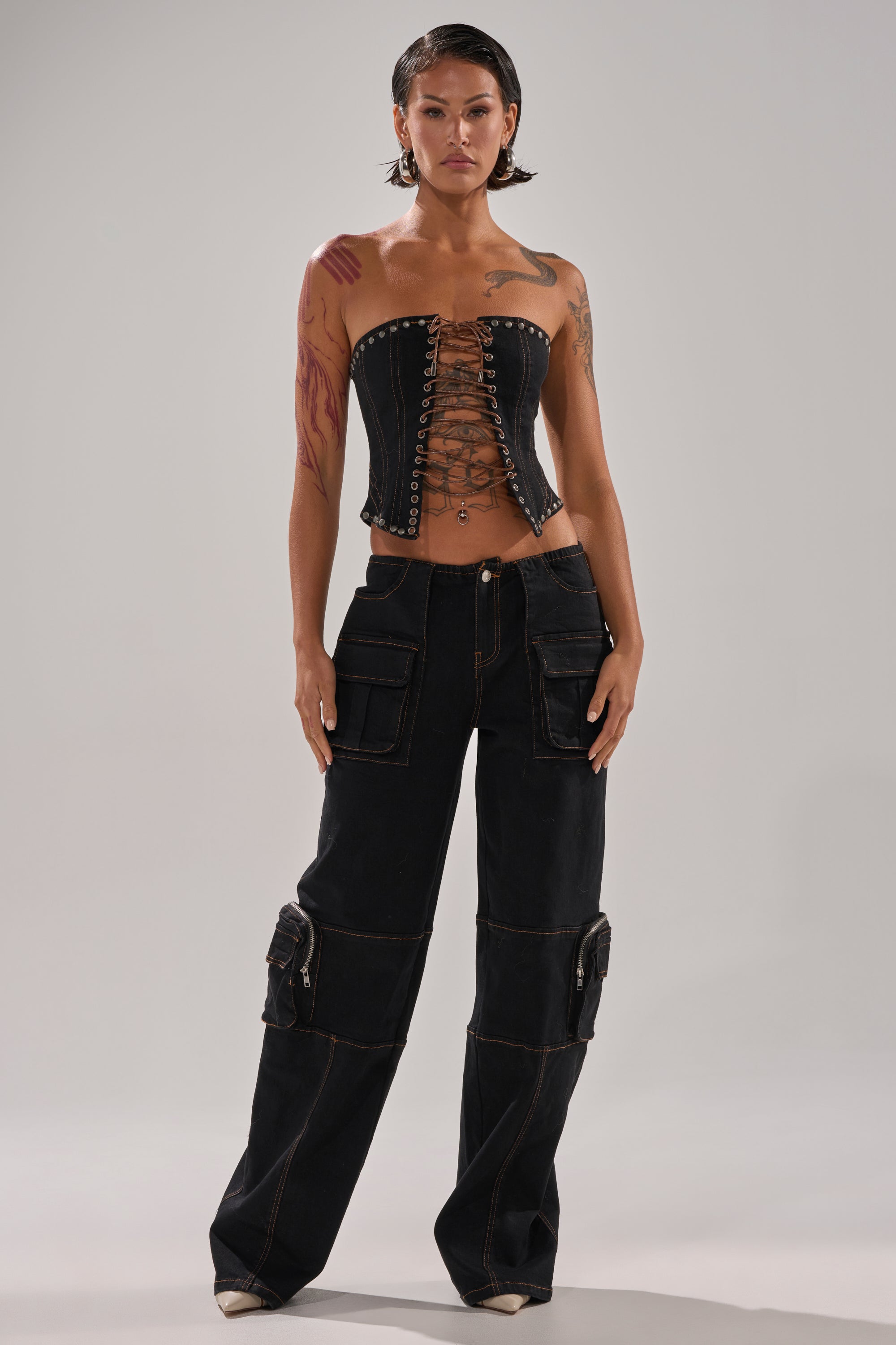 A person with short hair and tattoos stands confidently in a black lace-up corset top and the FEELING FRISKY CARGO DENIM PANT, featuring a wide-leg silhouette, large pockets, and visible stitching.