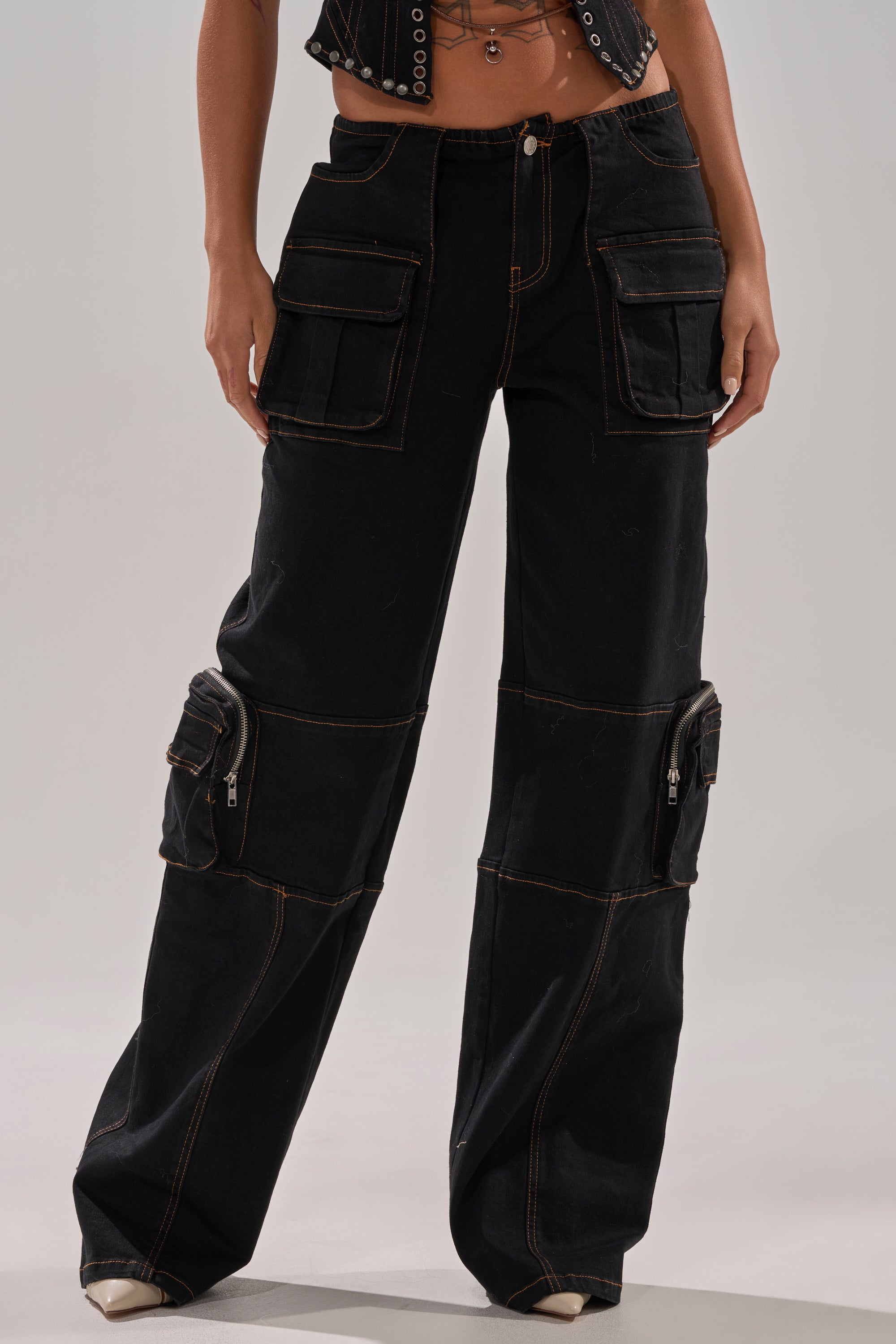 A person wears FEELING FRISKY CARGO DENIM PANT—high-waisted, black, wide-leg with brown stitching—paired with a cropped black top. Only the torso and legs are shown.